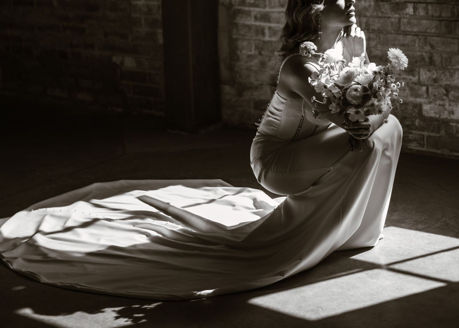 Wedding dress and bouquet create elegant shadows on sunlit floor in rustic brick setting.