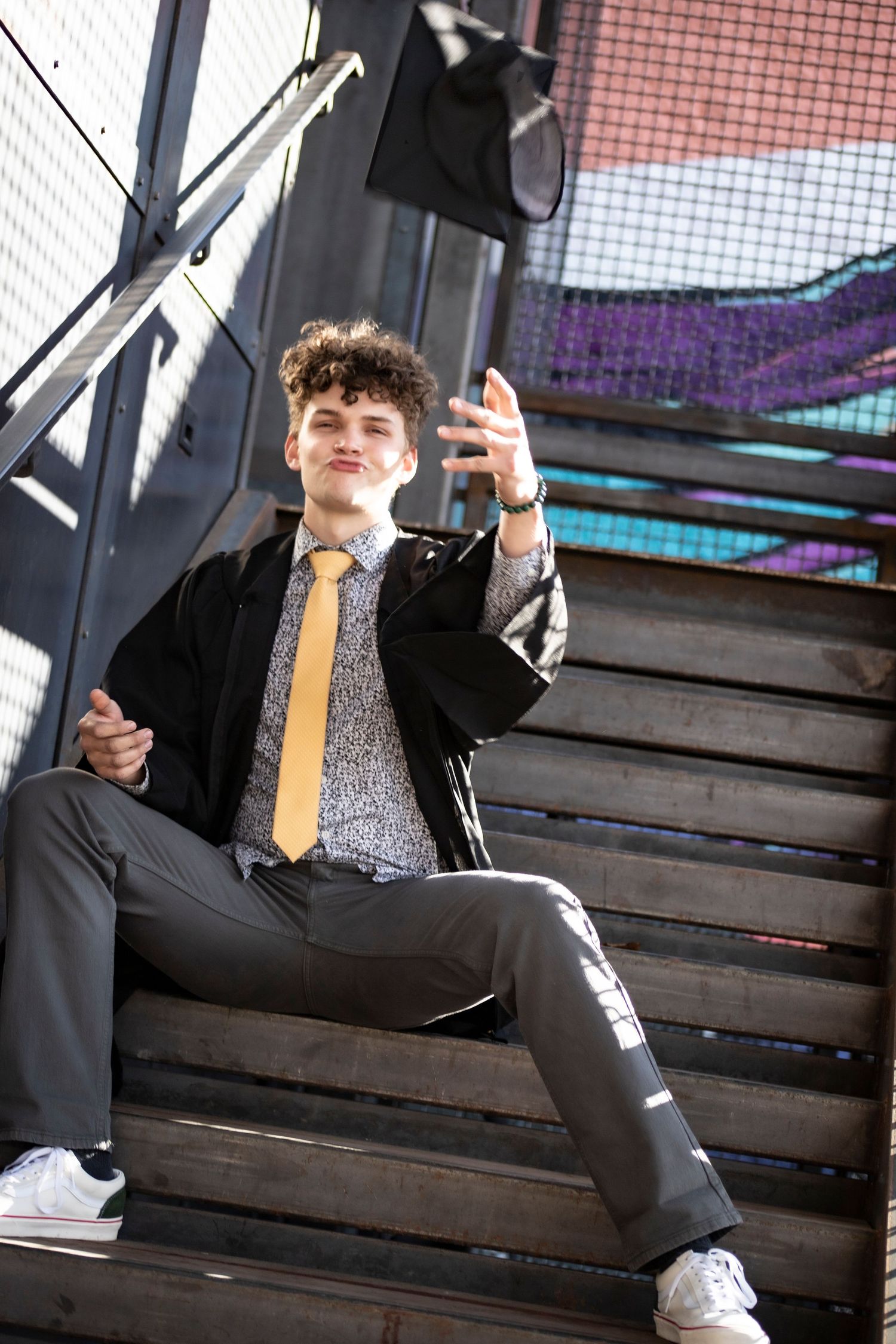 Graduate in cap and gown poses on industrial staircase with colorful backdrop.