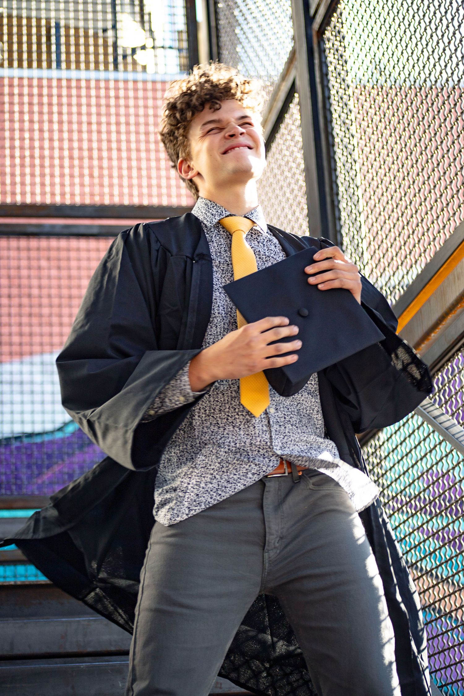Graduate holding diploma poses joyfully on metal staircase with urban background.