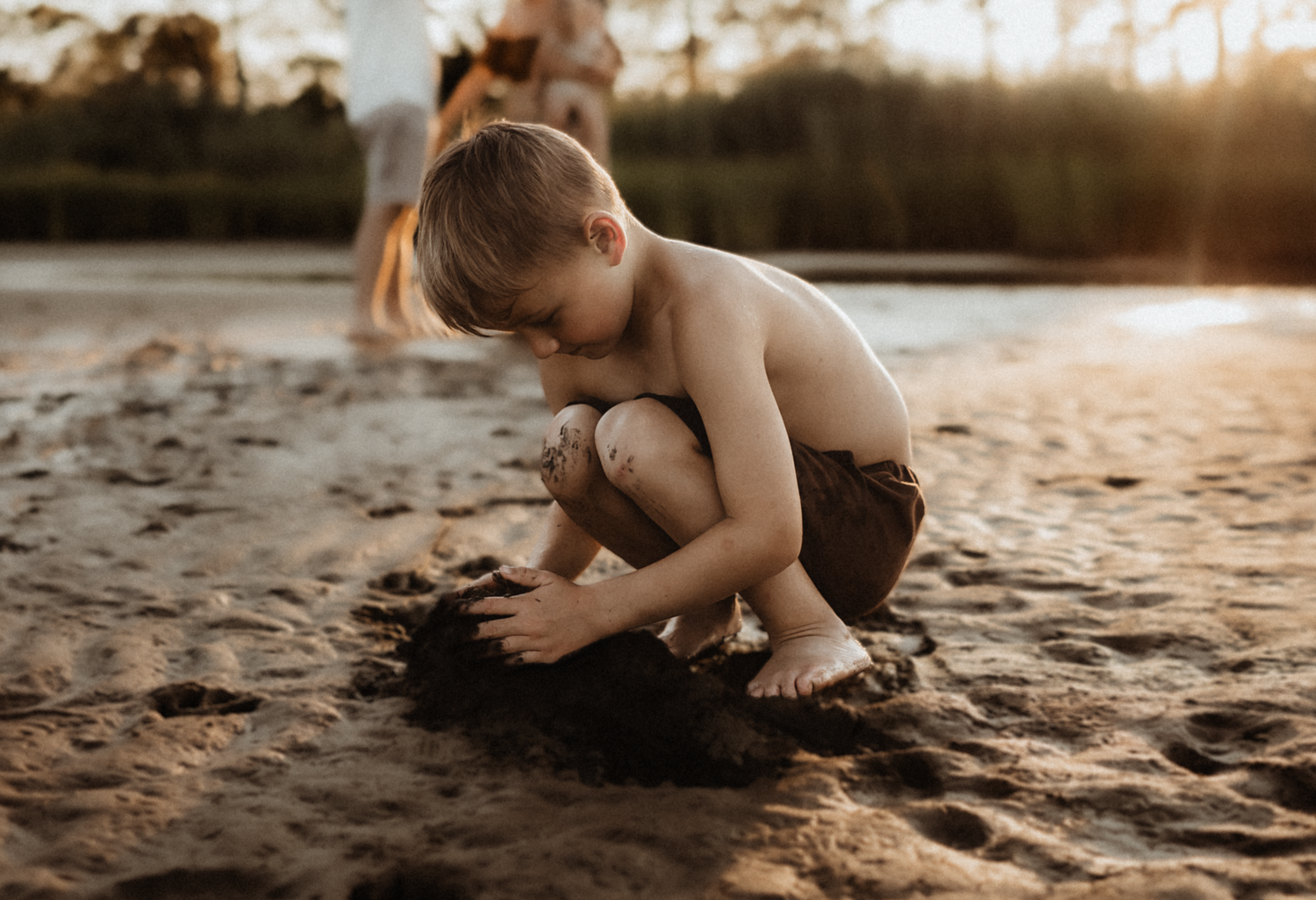 Shirtless young boy playing in wet sand on a beach at golden sunset hour, building sandcastles barefoot.