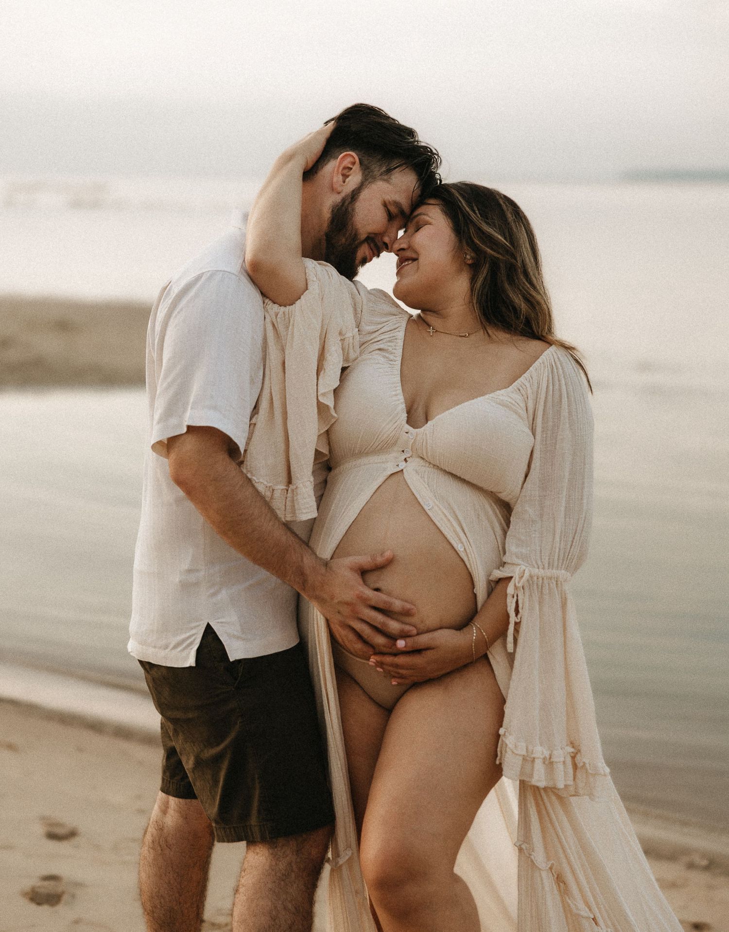 A couple shares intimate moments on a beach in light colored clothing during a maternity photoshoot at sunset.