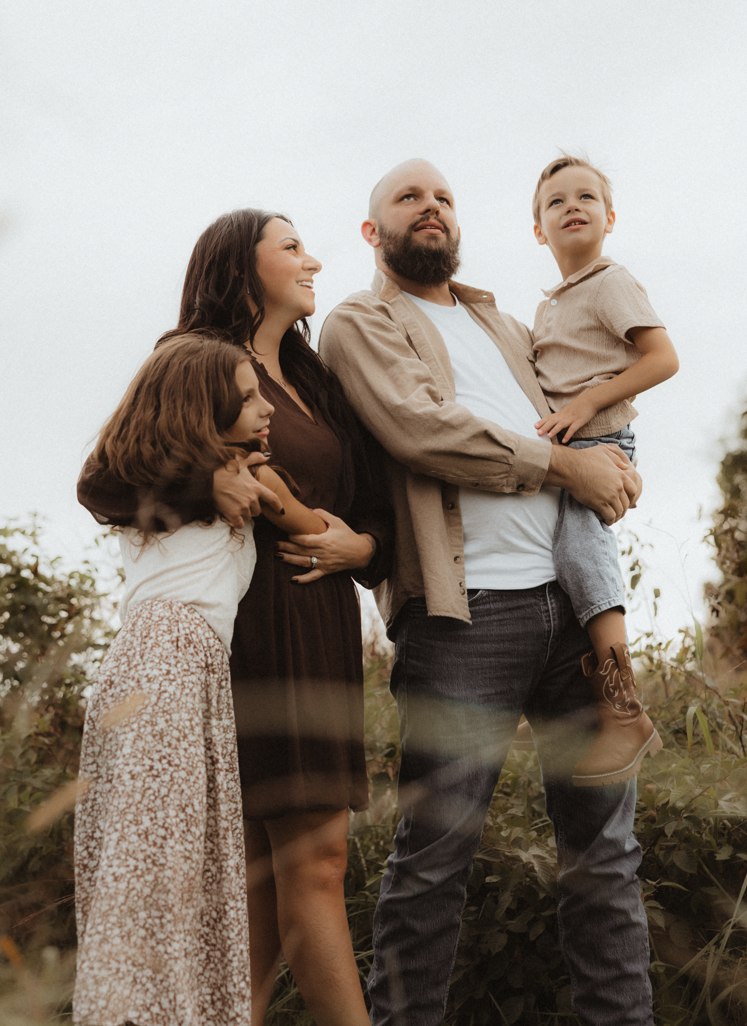 A family of four poses outdoors, parents holding children, dressed in neutral tones against a natural backdrop.