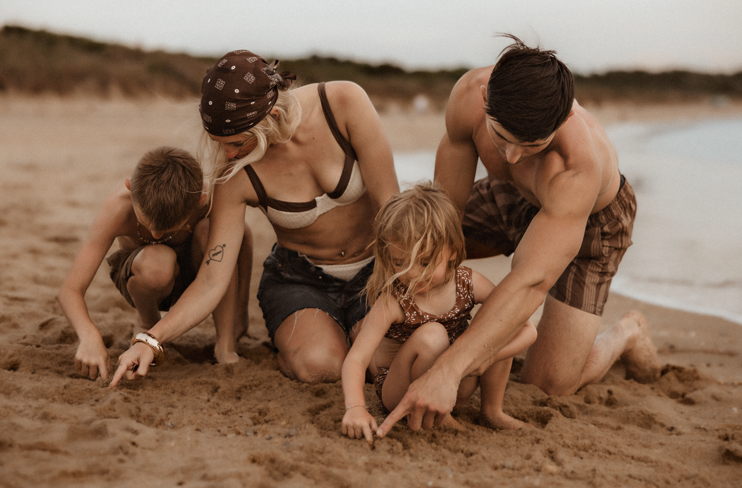 Family of four playing together in the sand on a beach at sunset, laughing and digging joyfully.