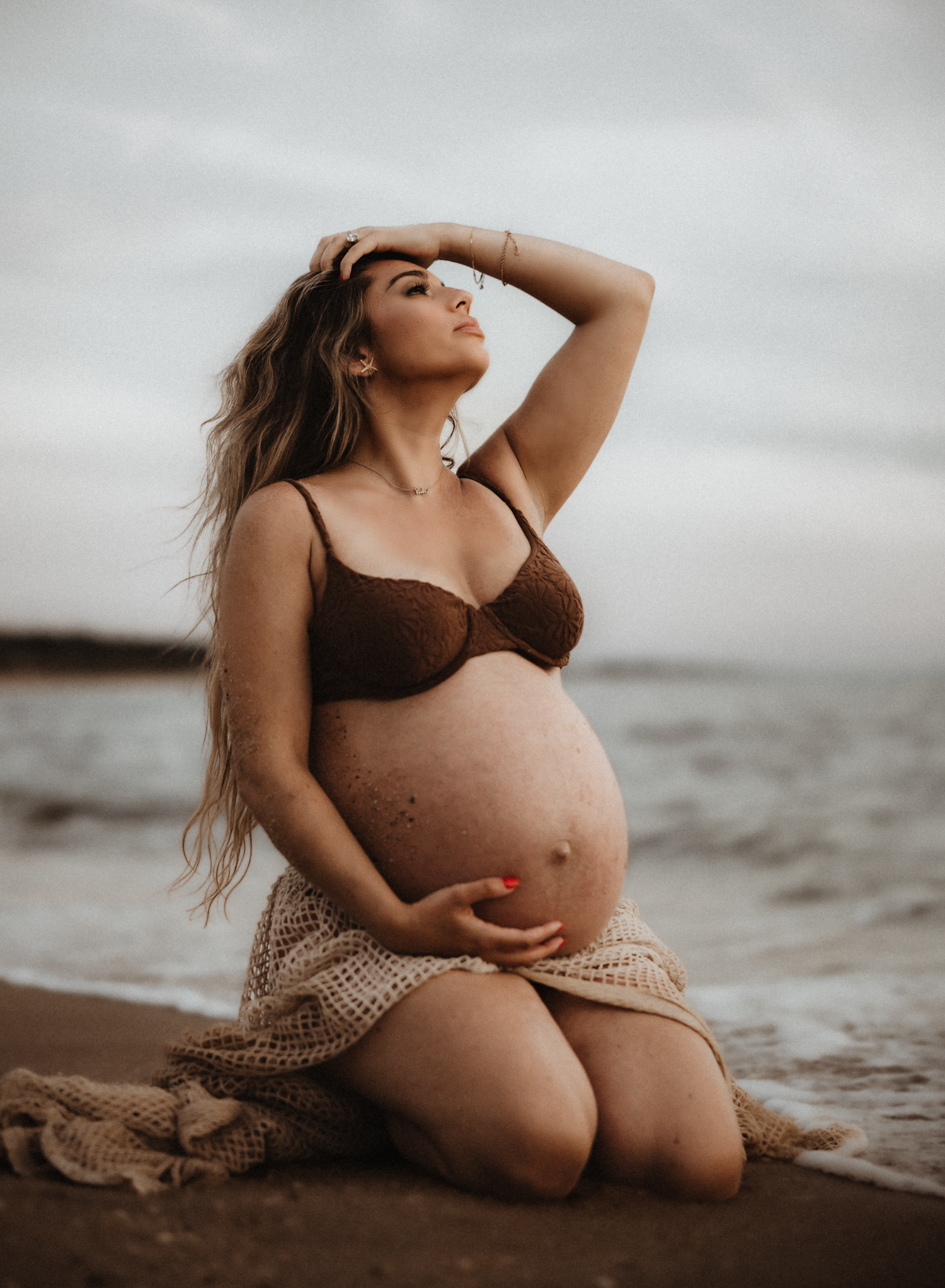 Pregnant woman in brown bikini kneeling on beach, cradling baby bump, with long blonde hair and ocean backdrop.