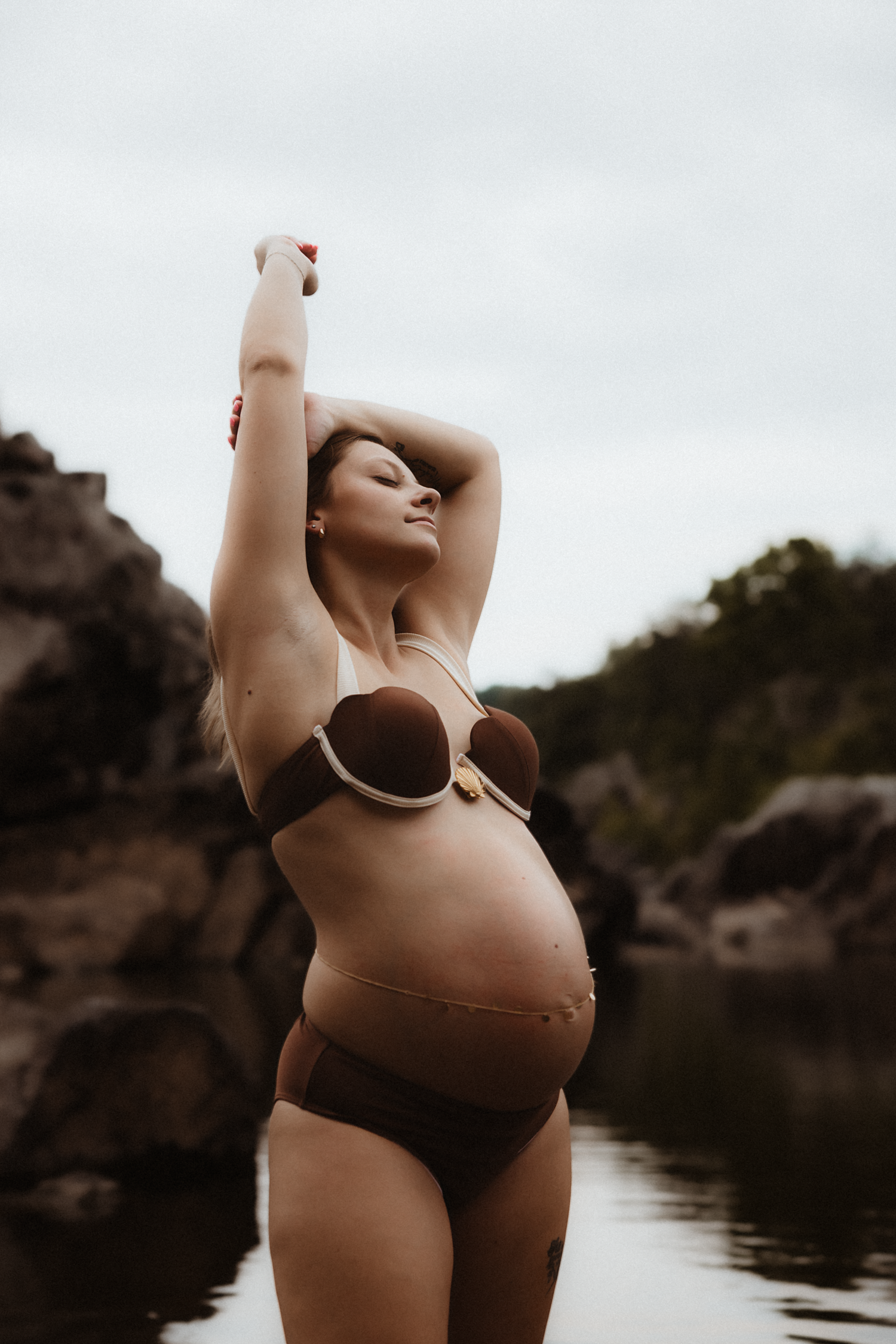 Pregnant woman in brown bikini poses with arms raised near rocky waterfront, showcasing baby bump outdoors.