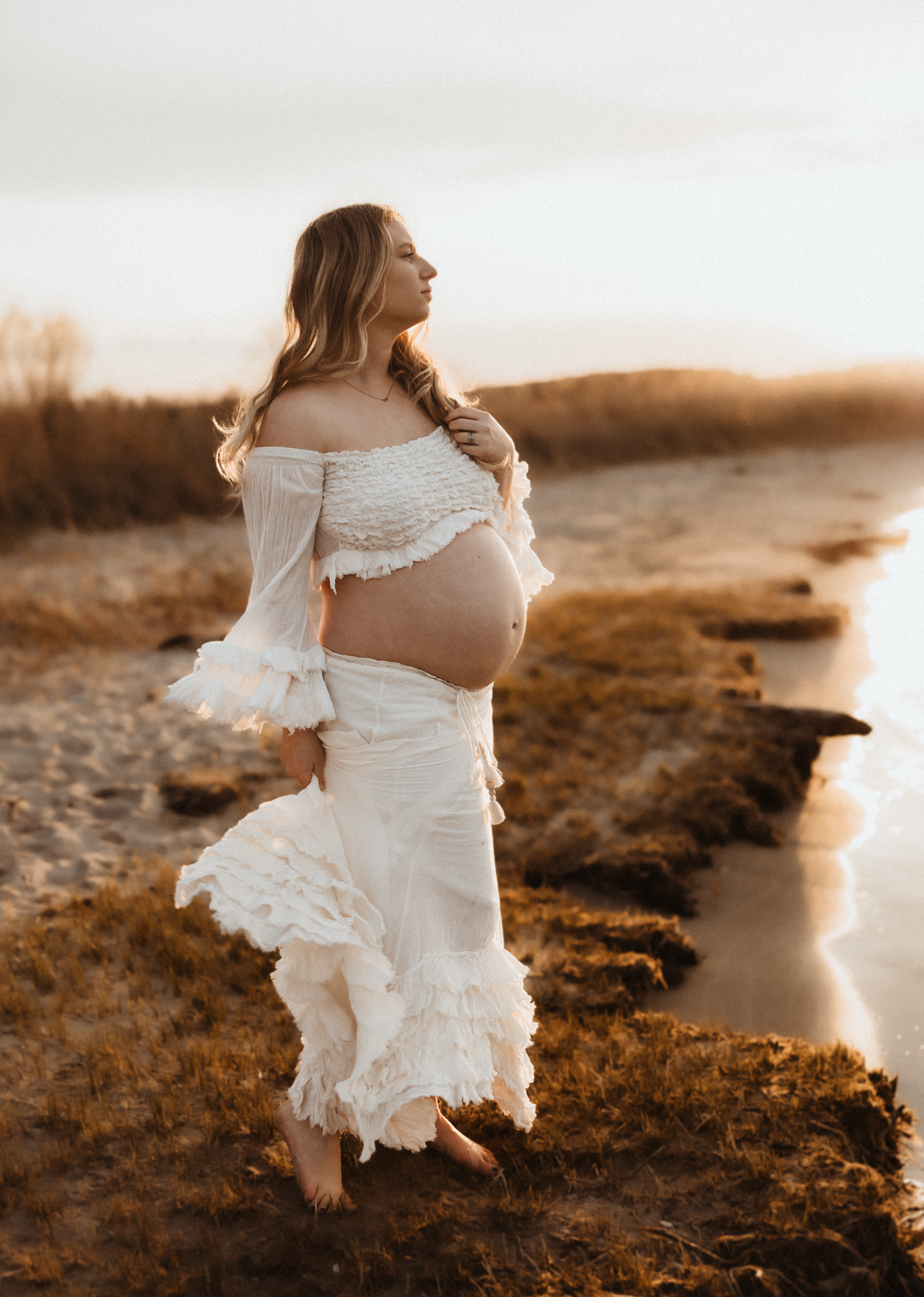 Pregnant woman in white off-shoulder dress poses gracefully on sandy shore at golden hour sunset.