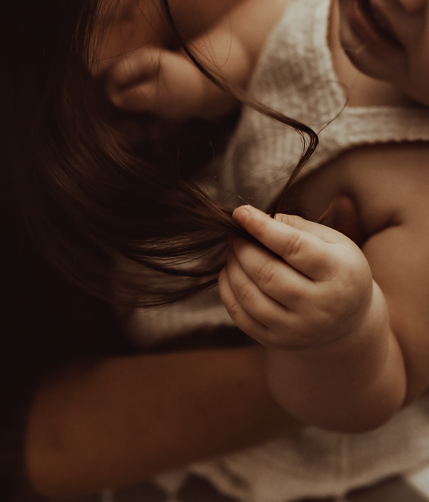 A toddler with dark hair gently holds a strand of hair between tiny fingers, wearing a light linen outfit.