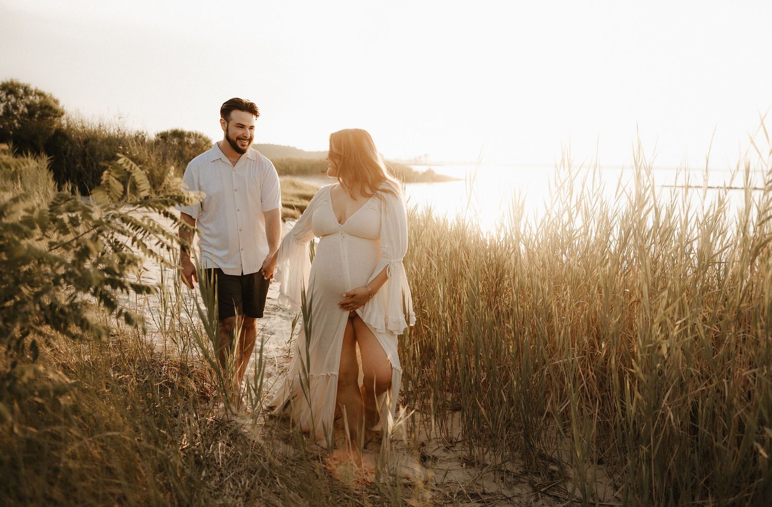 A couple in white outfits walks together through golden grass during sunset at a beach.