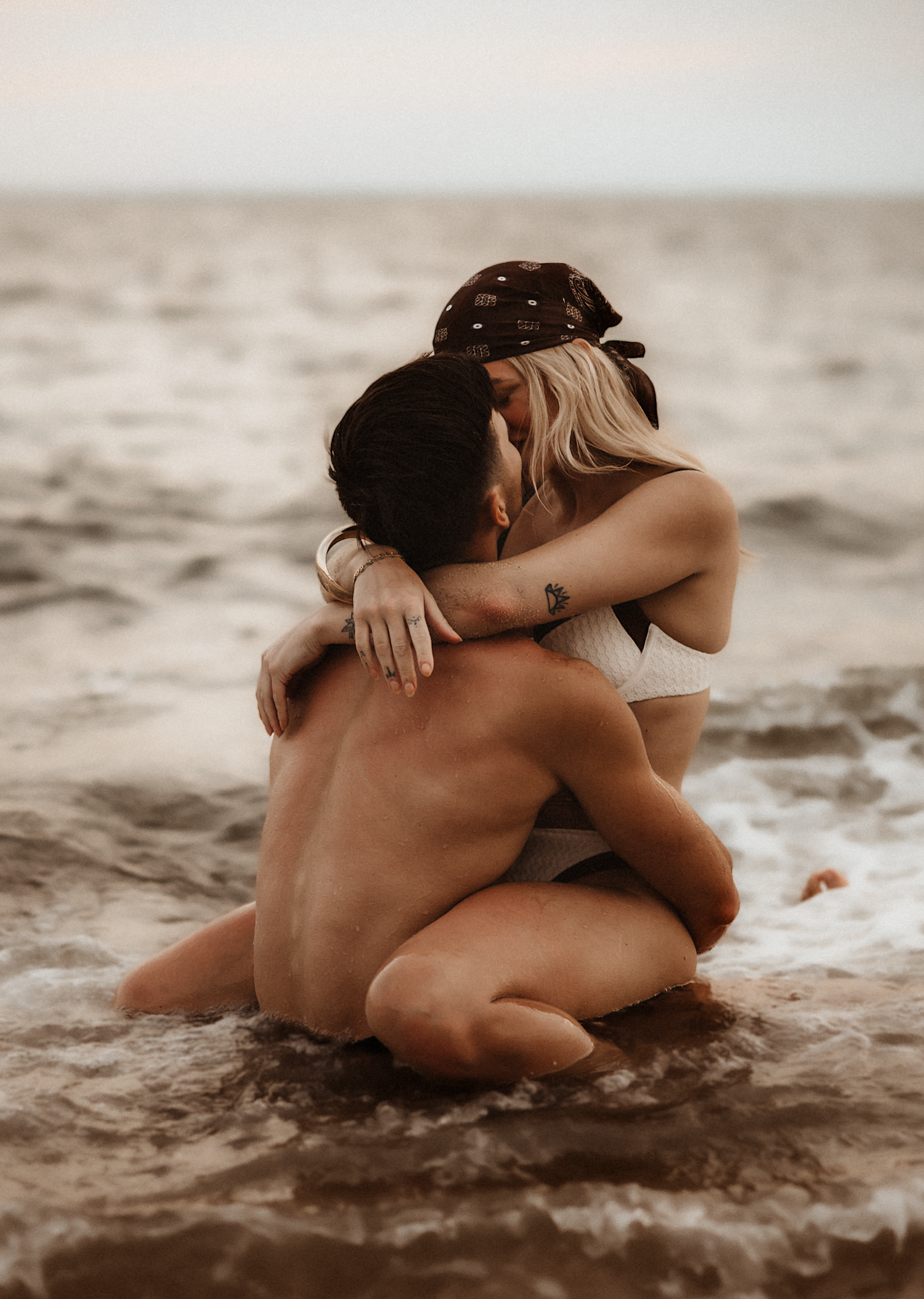 Couple embracing romantically in ocean waves on a beach, sitting in shallow water in a tender intimate moment.