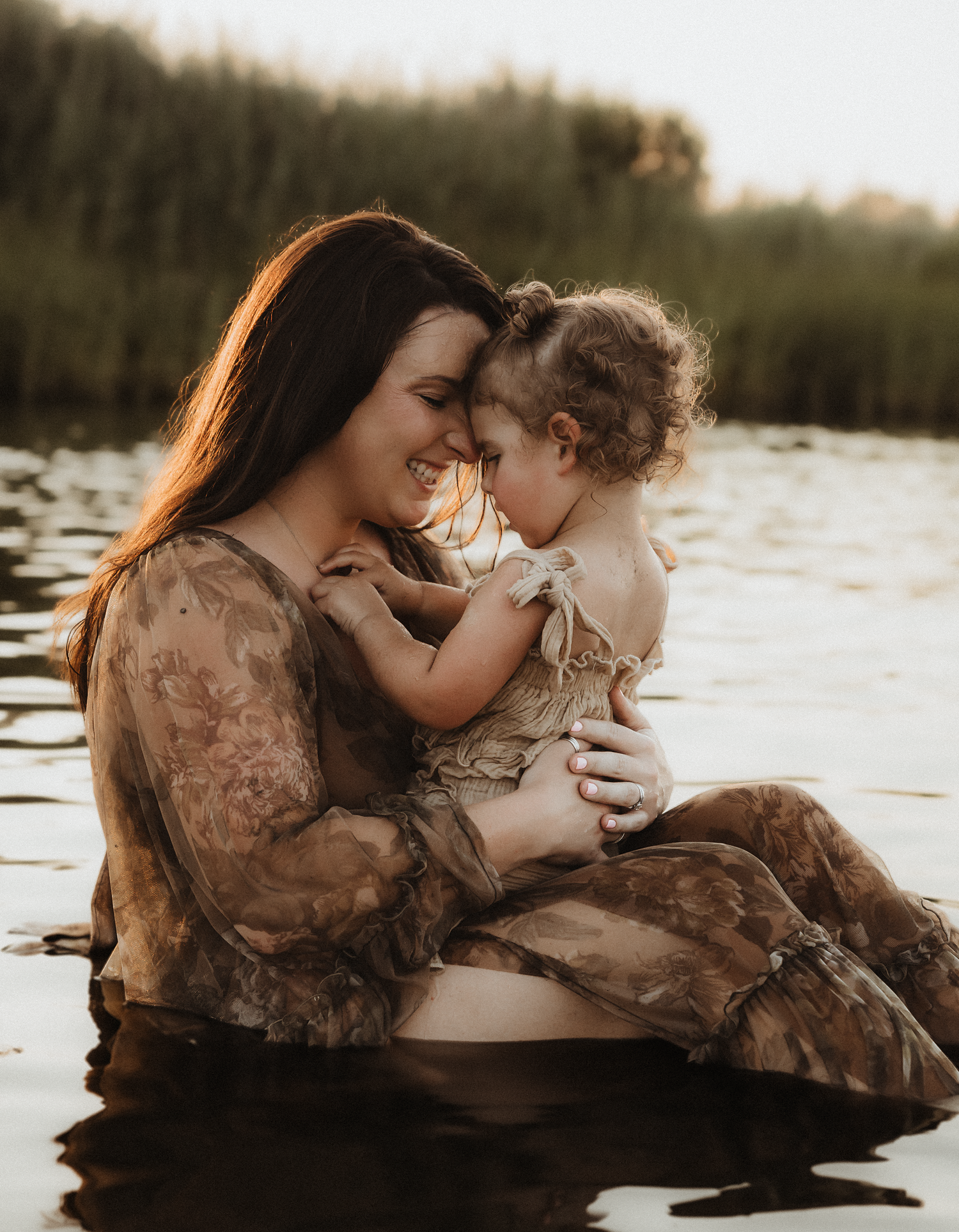 A mother smiles tenderly holding her toddler while sitting in a calm lake at golden hour.