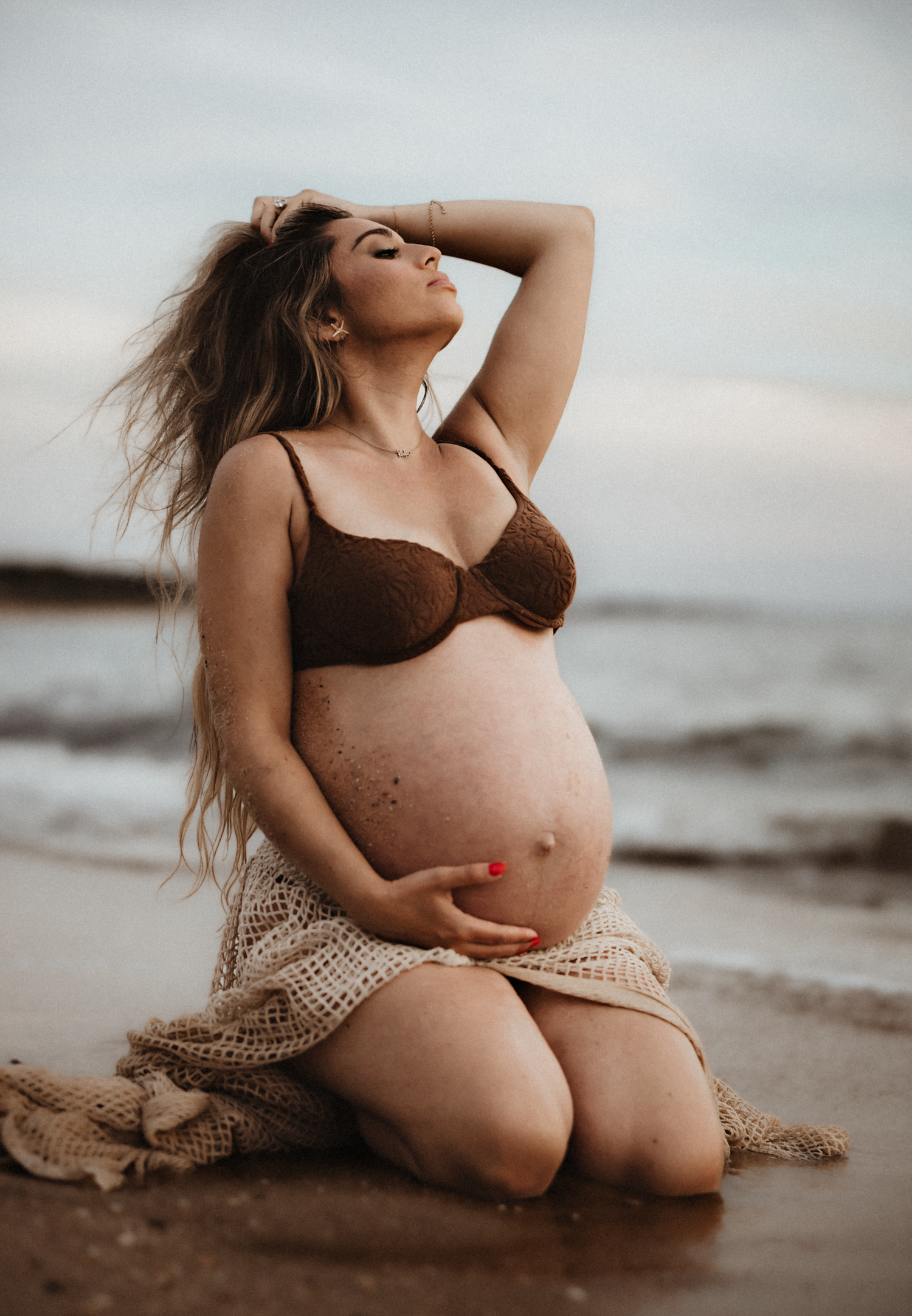 Pregnant woman kneeling on beach in brown bikini top and crochet skirt, cradling baby bump with ocean waves behind her.