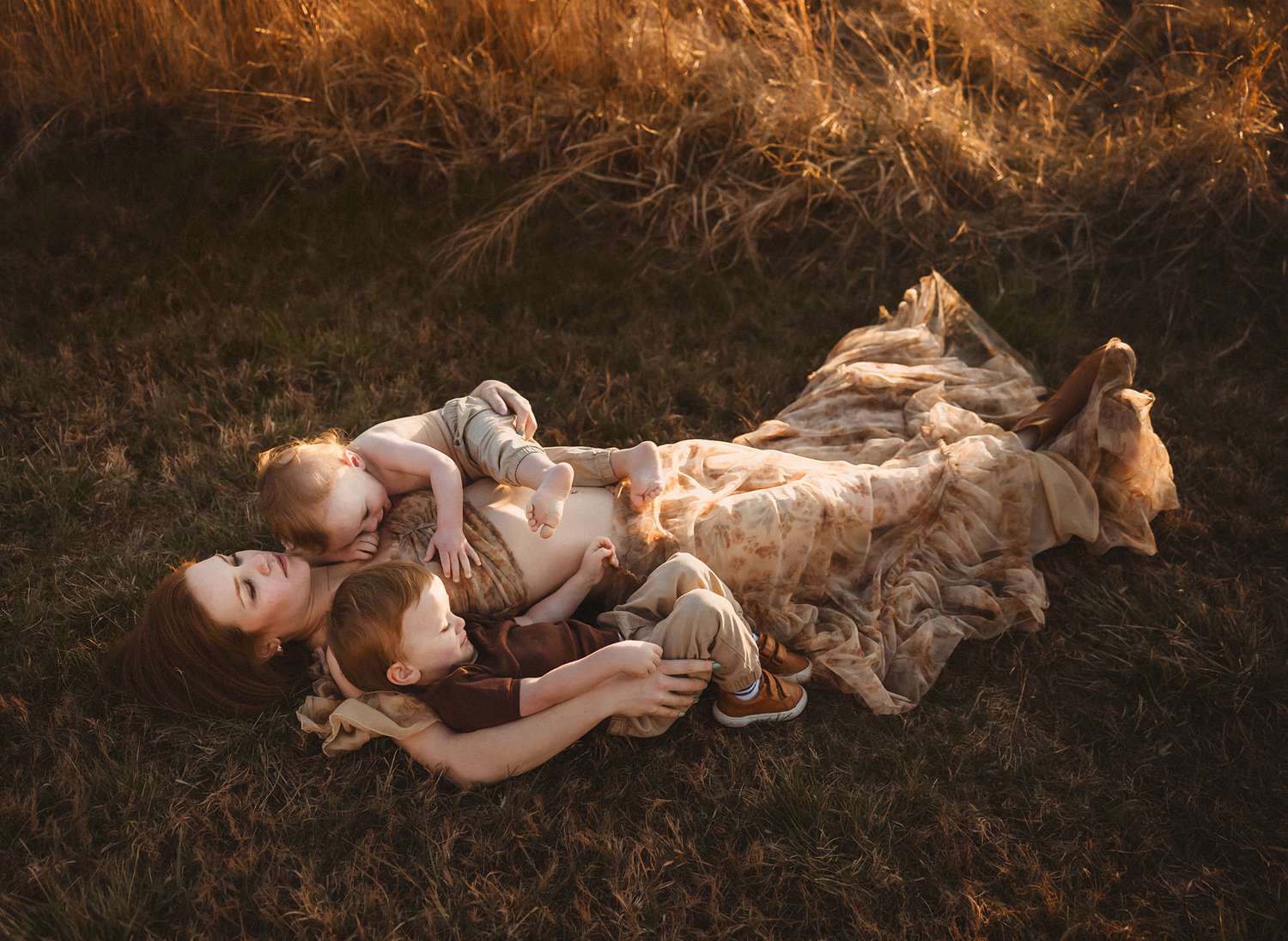 A mother lies in golden grass at sunset, cuddling two young children in a warm, tender outdoor portrait.