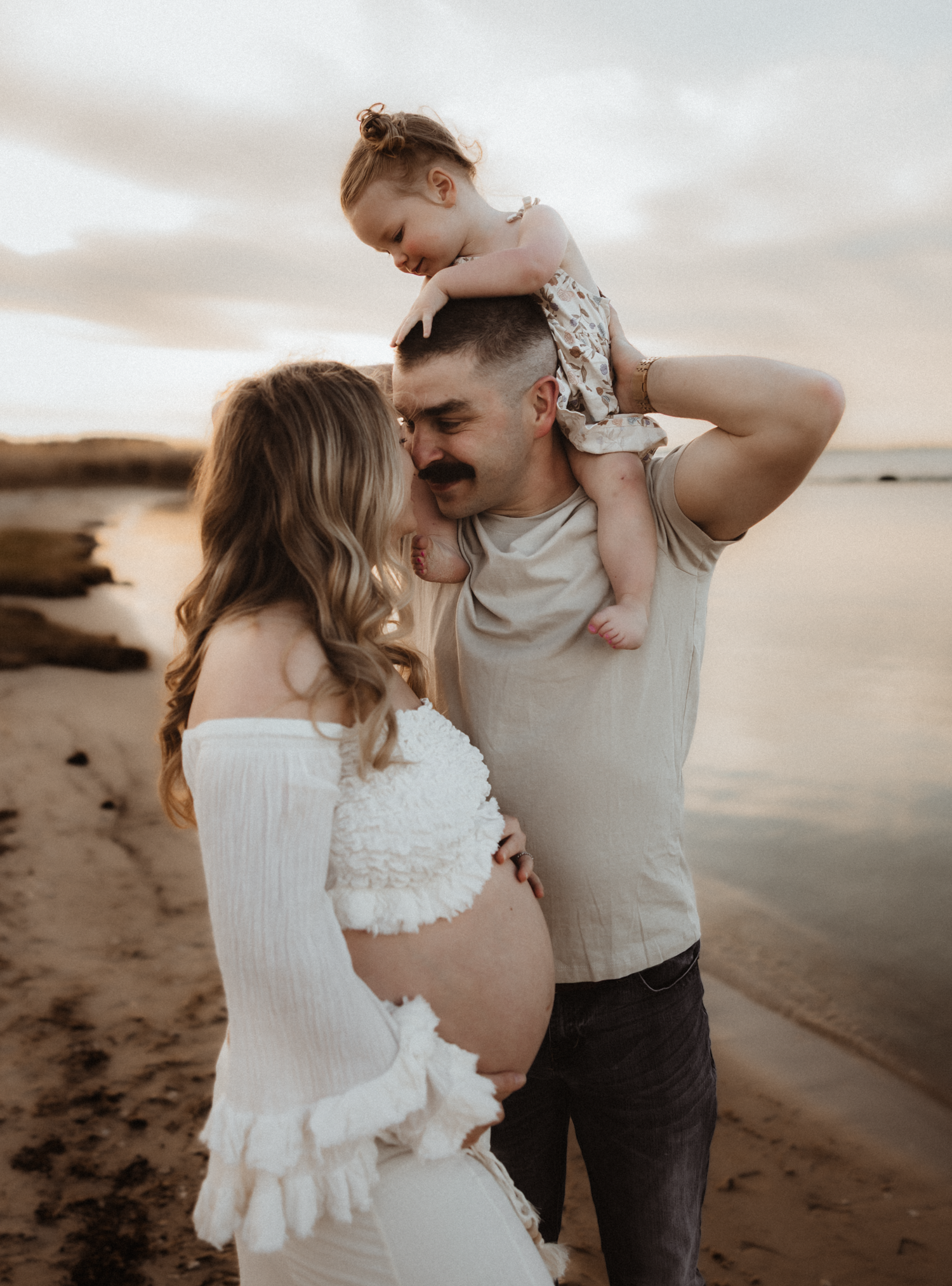 Pregnant woman in white dress with husband holding toddler on shoulders at beach during golden hour sunset.