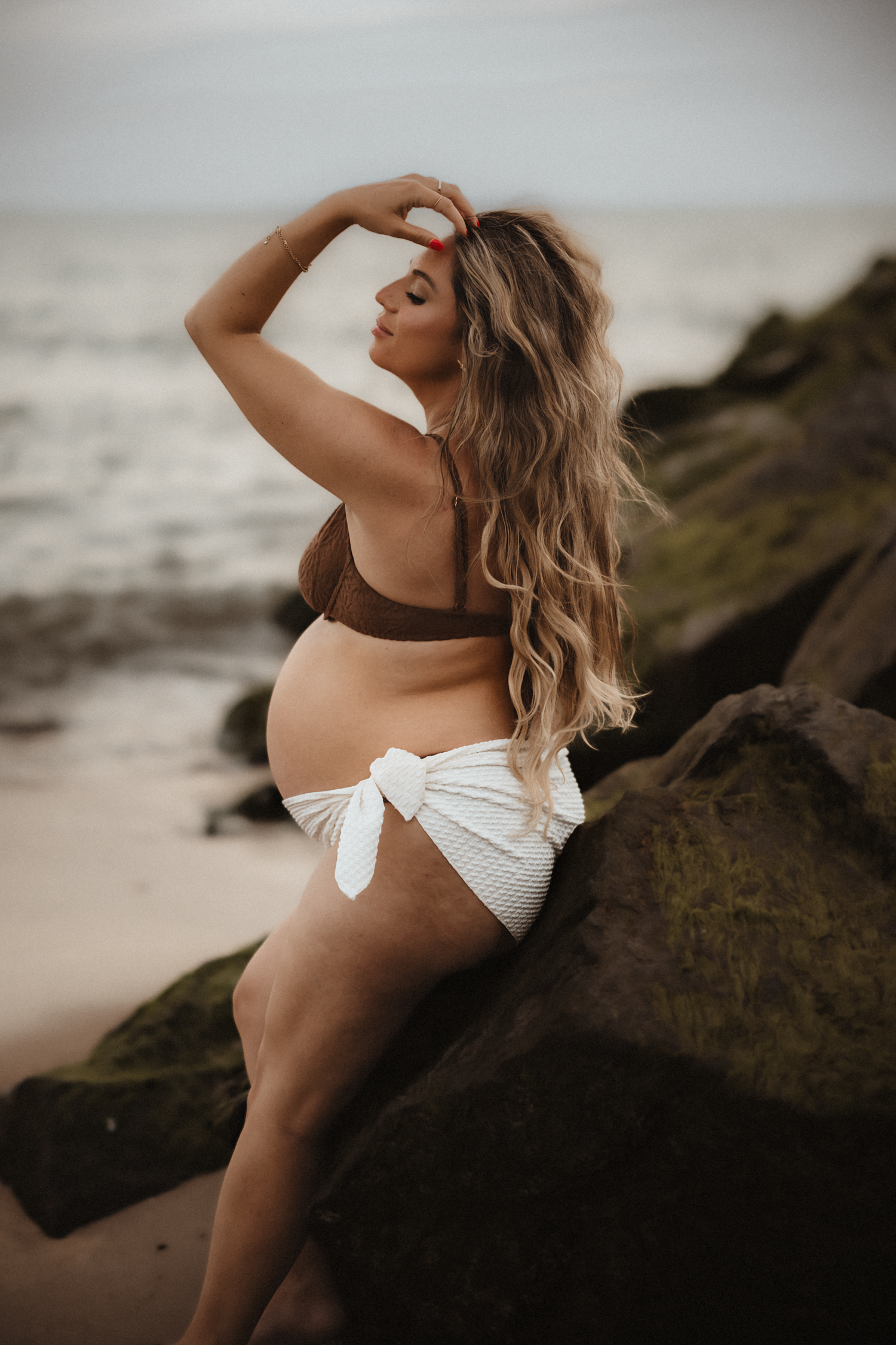 Pregnant woman in brown bikini top and white wrap bottom poses on mossy rocks at a beach.
