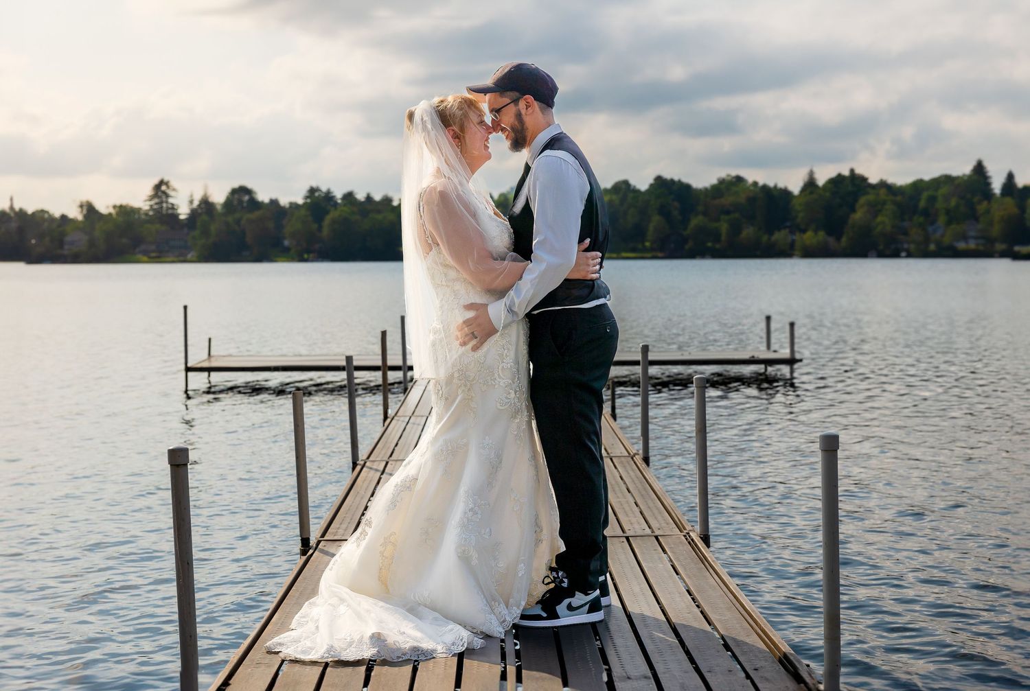 Wedding couple embracing on wooden dock with lake and sunset in background.