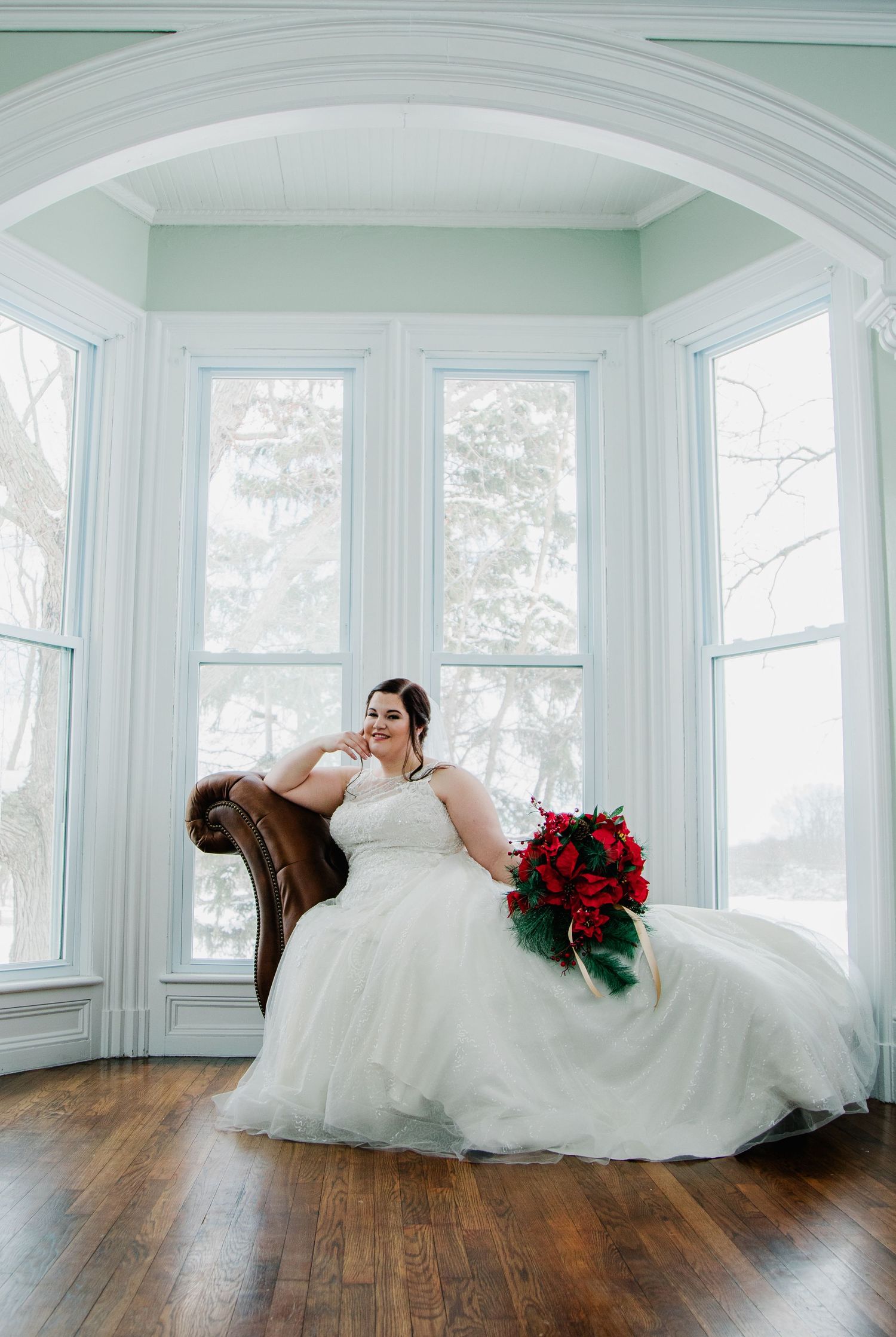 A bride in a white ballgown poses in an elegant bay window alcove with red roses and hardwood floors.