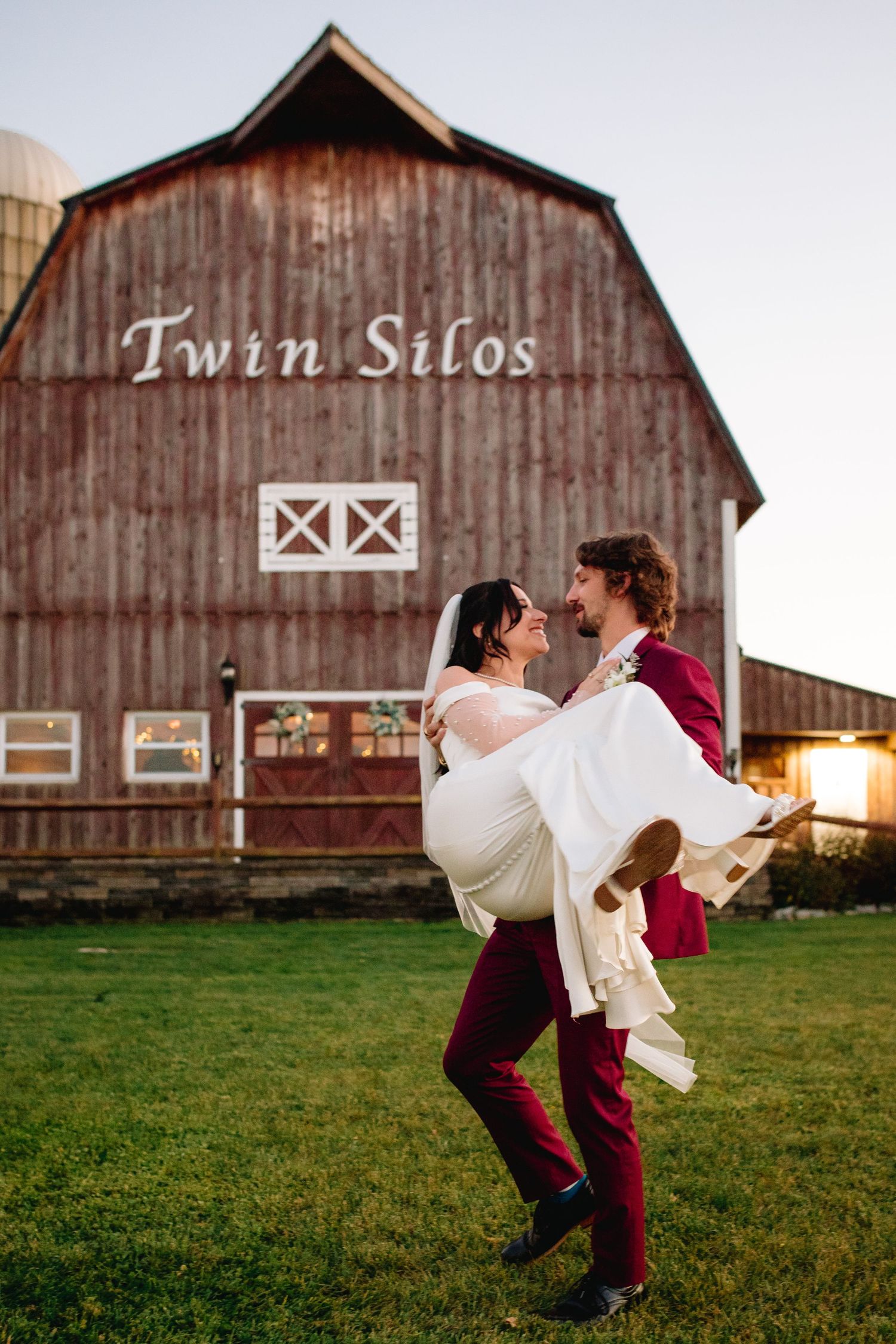 A romantic couple embraces in front of a rustic red barn with 'Twin Silos' text at sunset.