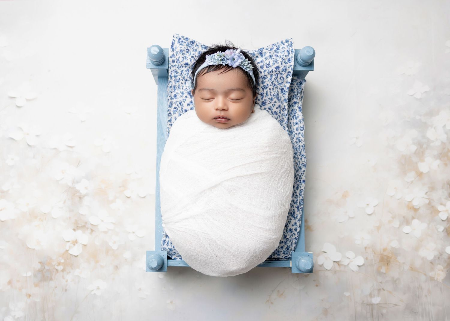 Newborn baby swaddled in white wrap sleeping peacefully in a small blue wooden bed with floral bedding.
