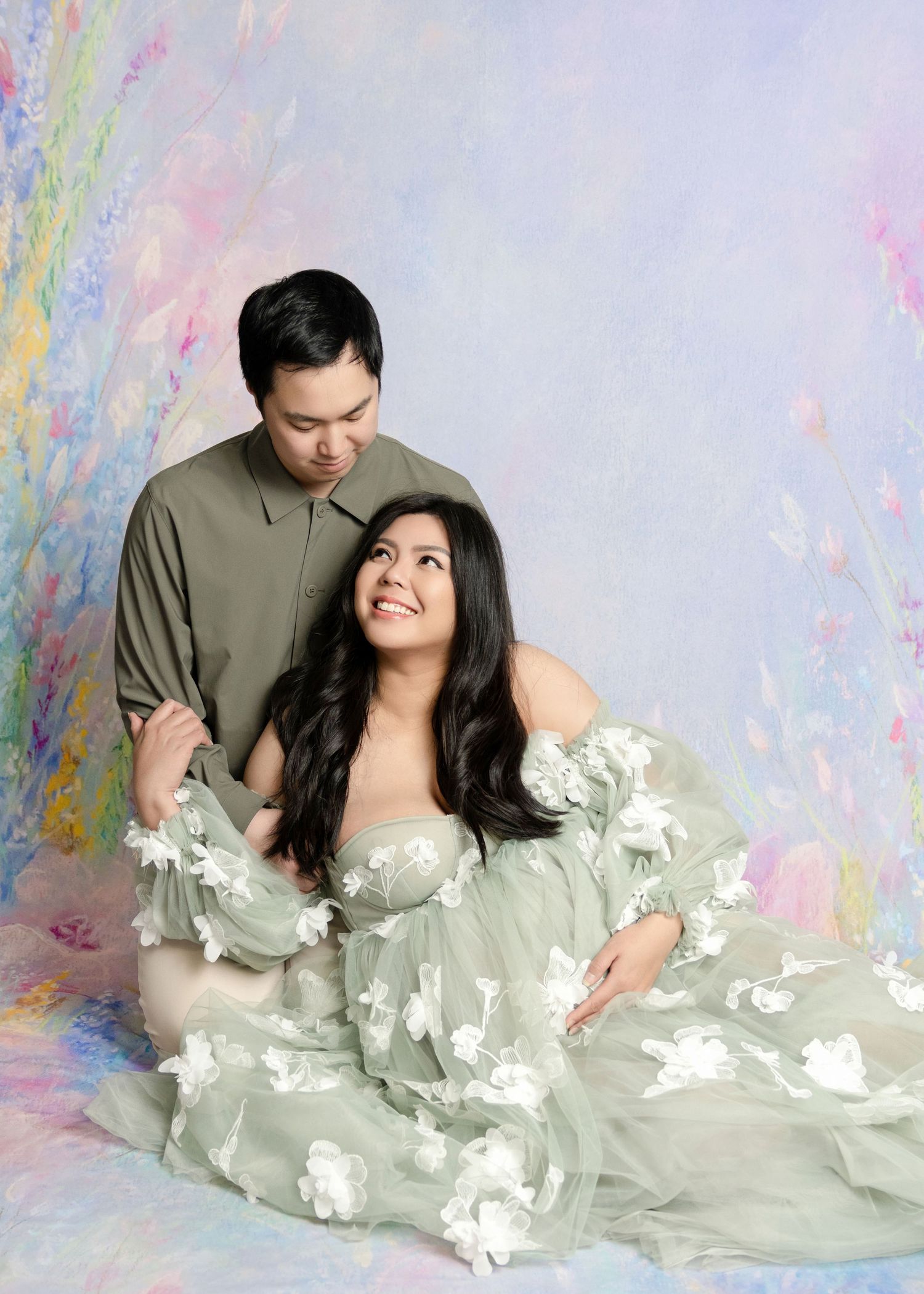 Couple in matching sage green outfits pose for a maternity photo shoot against a soft floral painted backdrop.