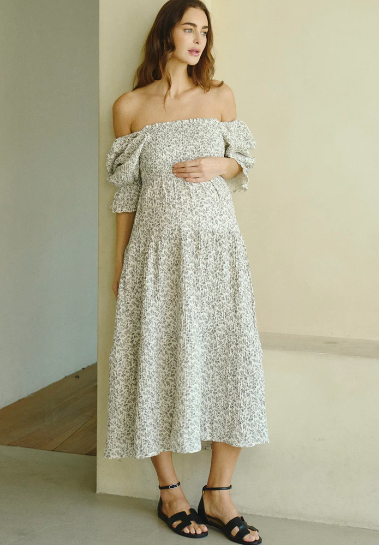 Pregnant woman wearing a white floral off-shoulder smocked midi dress with puff sleeves and black sandals.