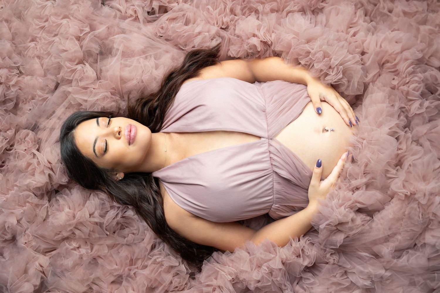 Pregnant woman lying on pink tulle fabric in a mauve wrap dress, cradling her baby bump with eyes closed.