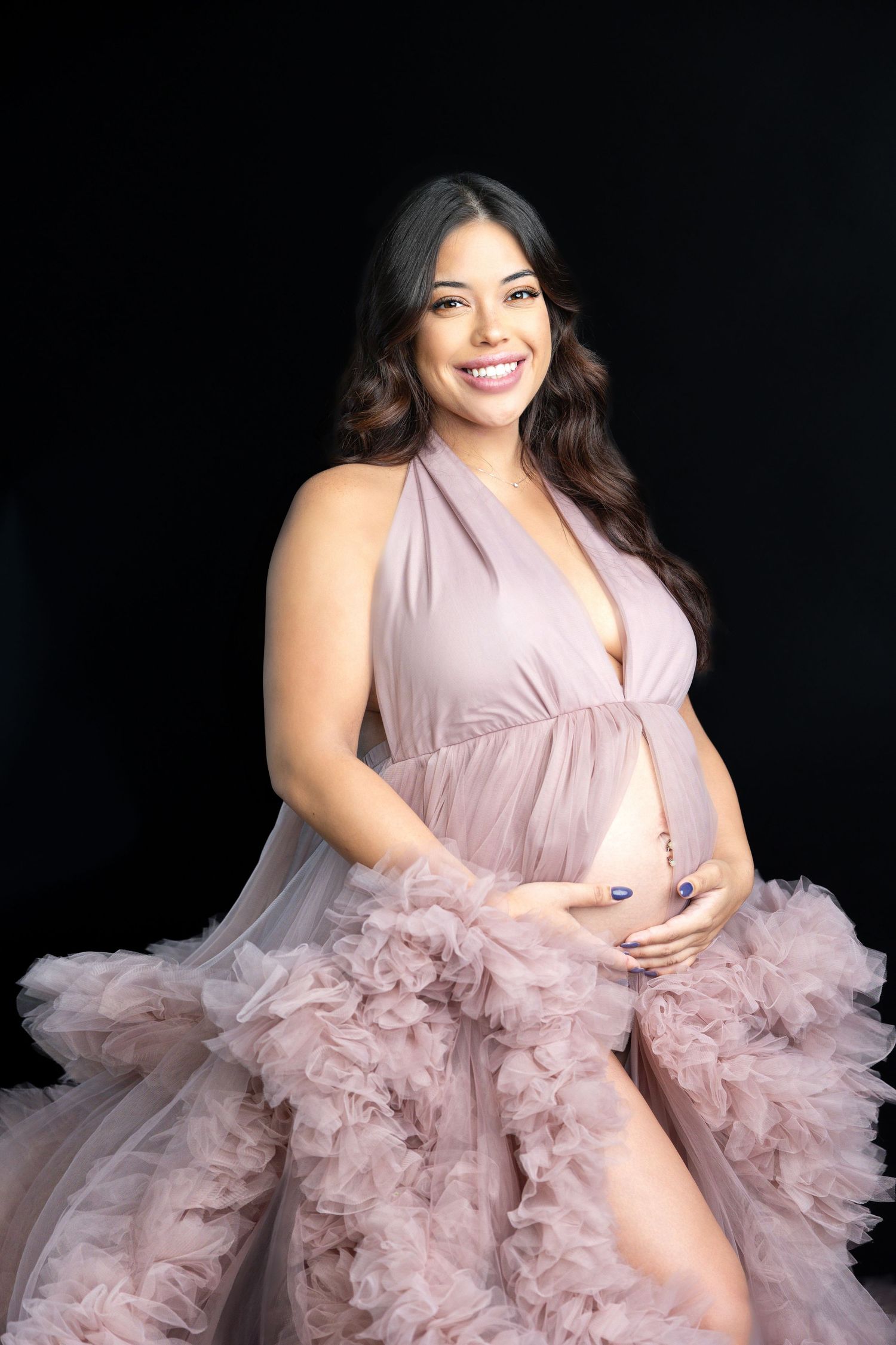 Smiling pregnant woman in pink ruffled maternity gown happily holding bare baby bump in studio.