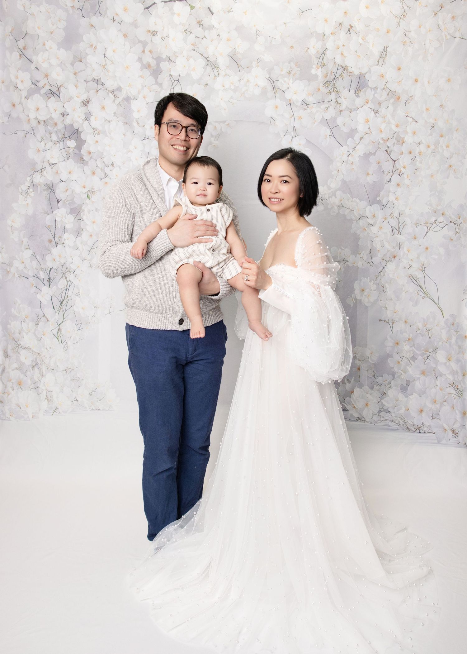 Asian family studio portrait with father, mother in white gown, and baby on floral backdrop.