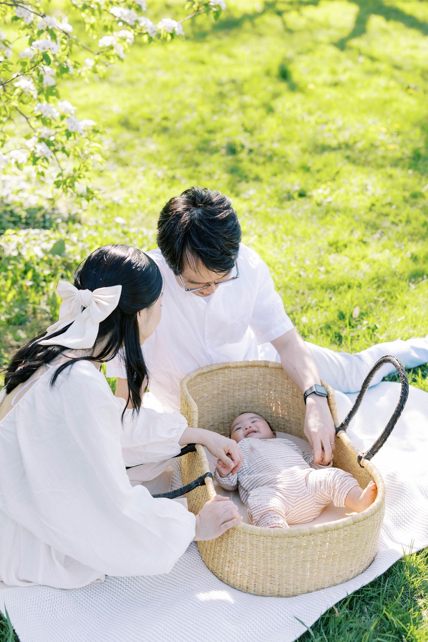 Young Asian parents lovingly tend to their newborn baby lying in a woven Moses basket on a sunny lawn.