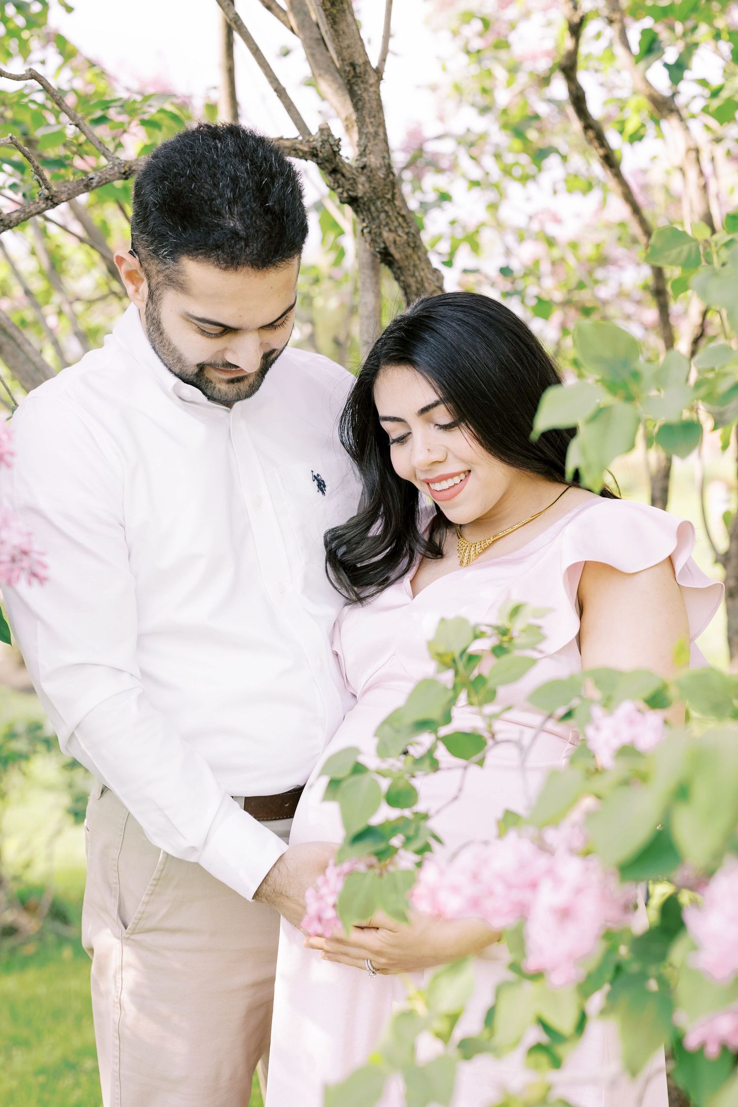 Expecting couple smiles tenderly while touching baby bump surrounded by pink blossoming trees.