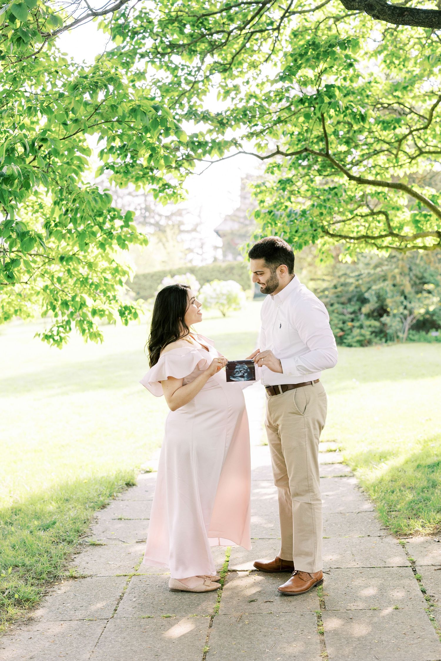 Expectant couple holding ultrasound photo on garden path under green trees, woman in pink dress.