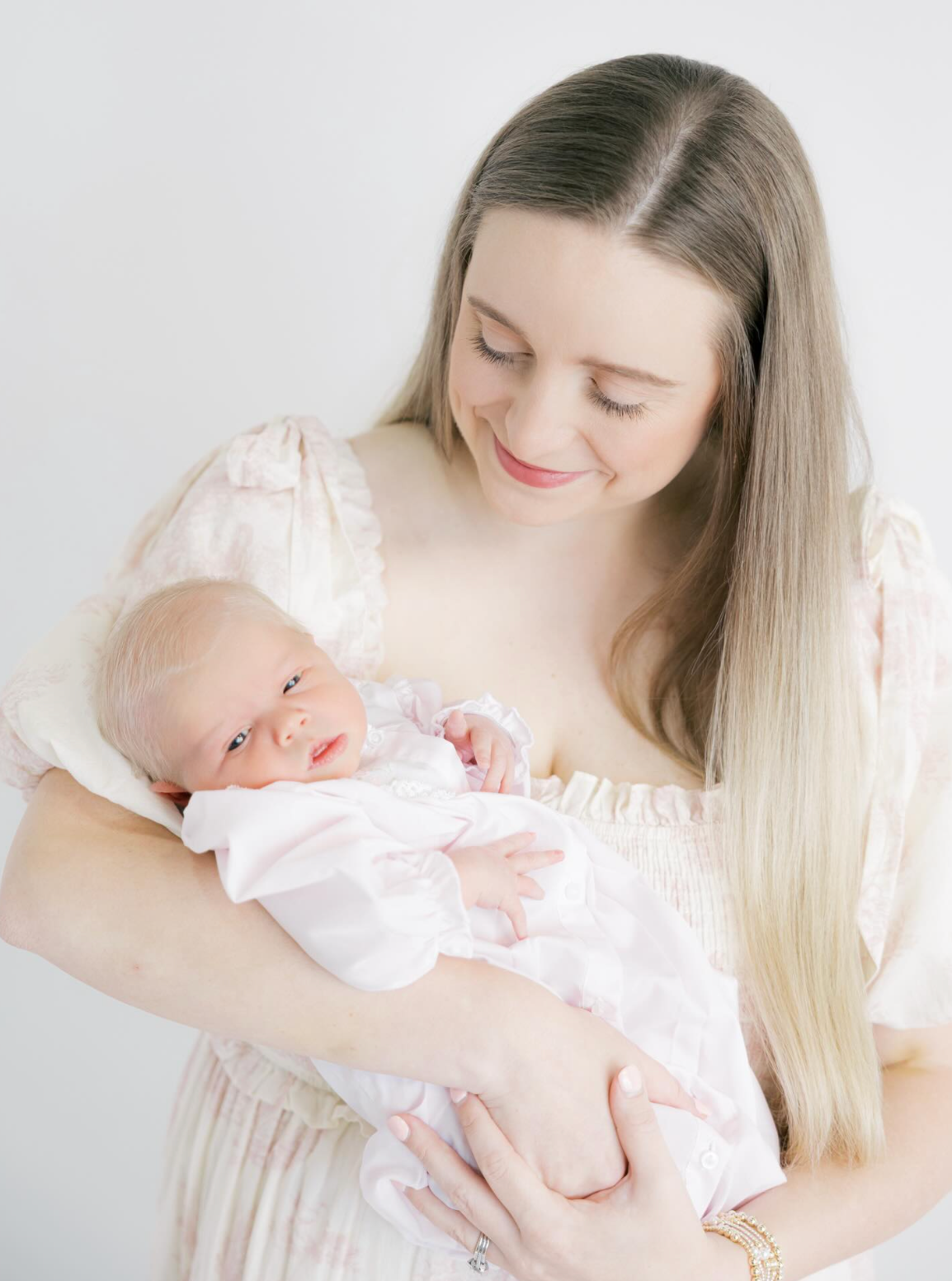 A smiling mother with long blonde hair holds her newborn baby in a soft pink outfit against a white background.