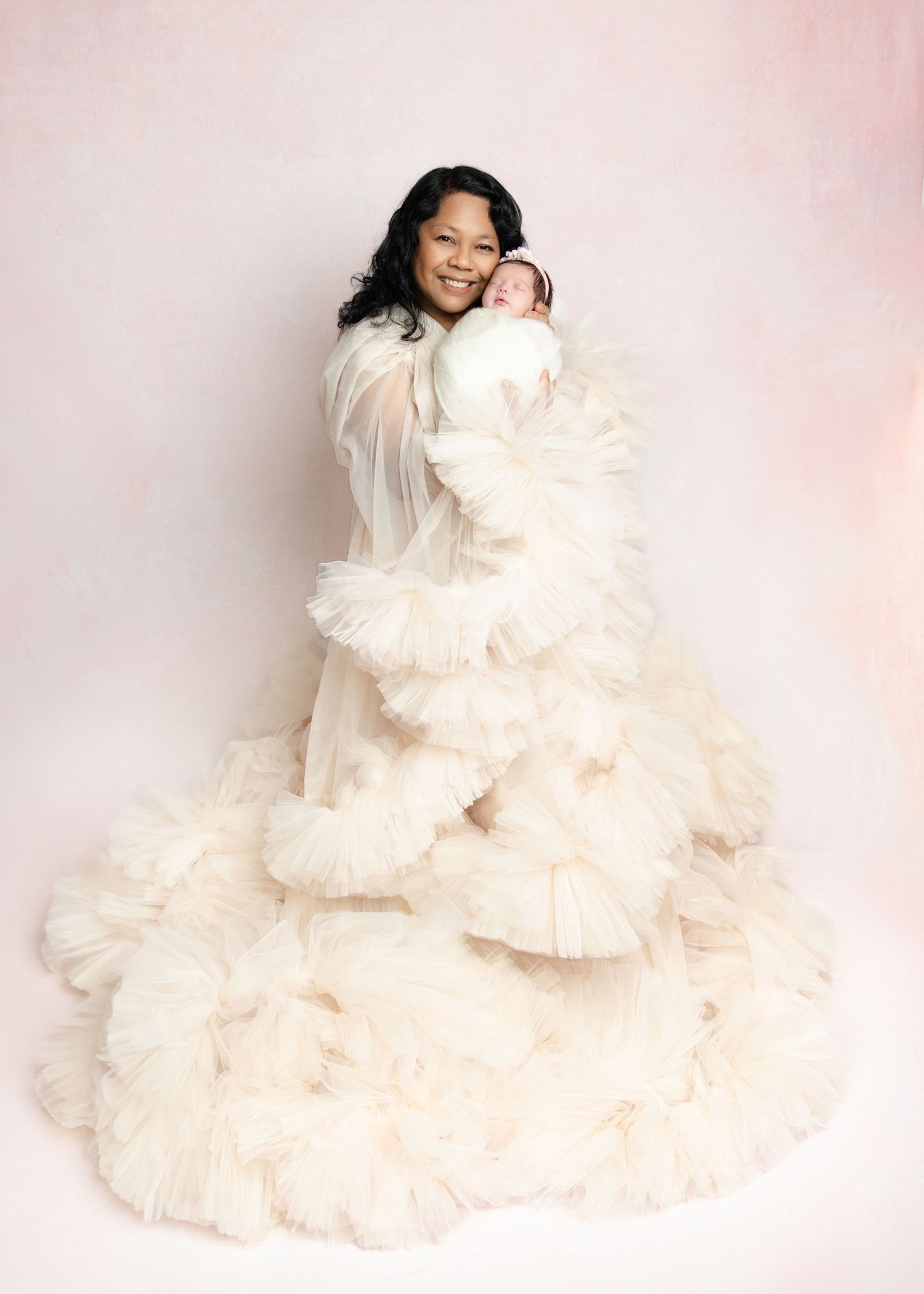 Woman in ivory ruffled tulle gown smiling while gently holding a sleeping newborn baby.