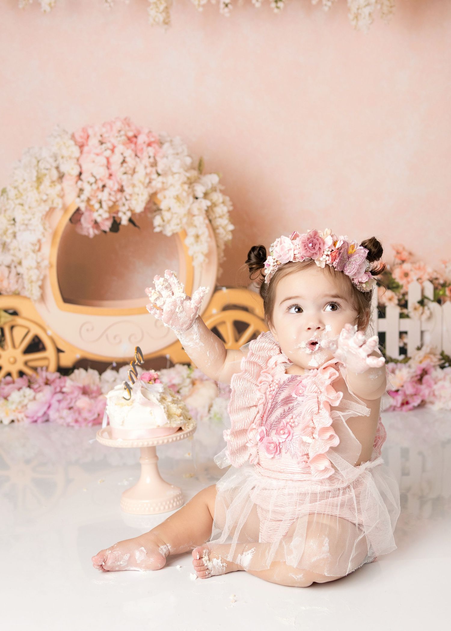 Baby girl in pink ruffle outfit with flower crown reaches up during princess themed first birthday cake smash.