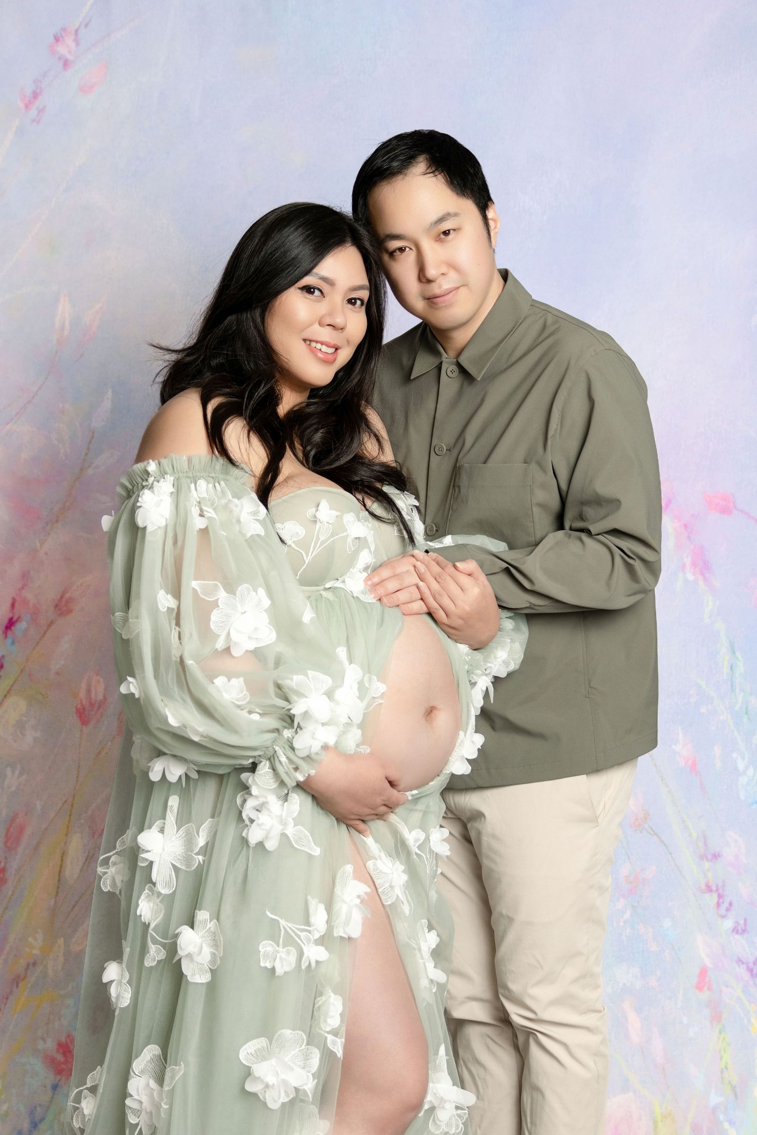 Couple posing for a maternity photo shoot, woman in floral green gown cradling baby bump, man in olive shirt embracing her.