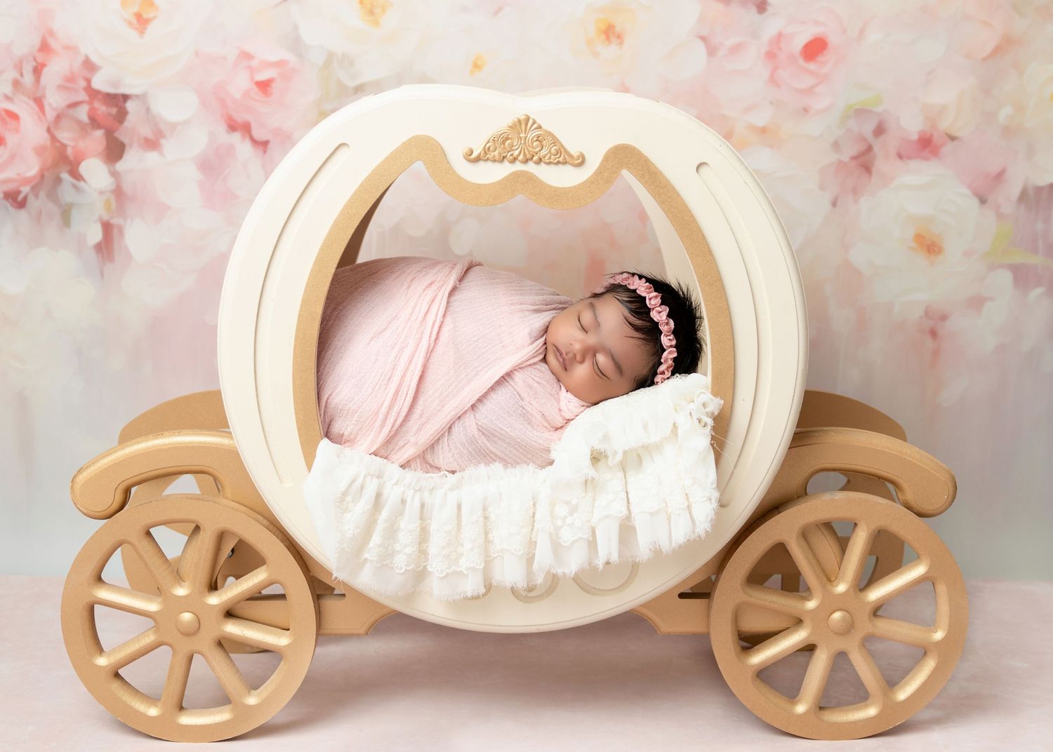Newborn baby sleeping peacefully in a white and gold Cinderella carriage prop with pink floral backdrop.