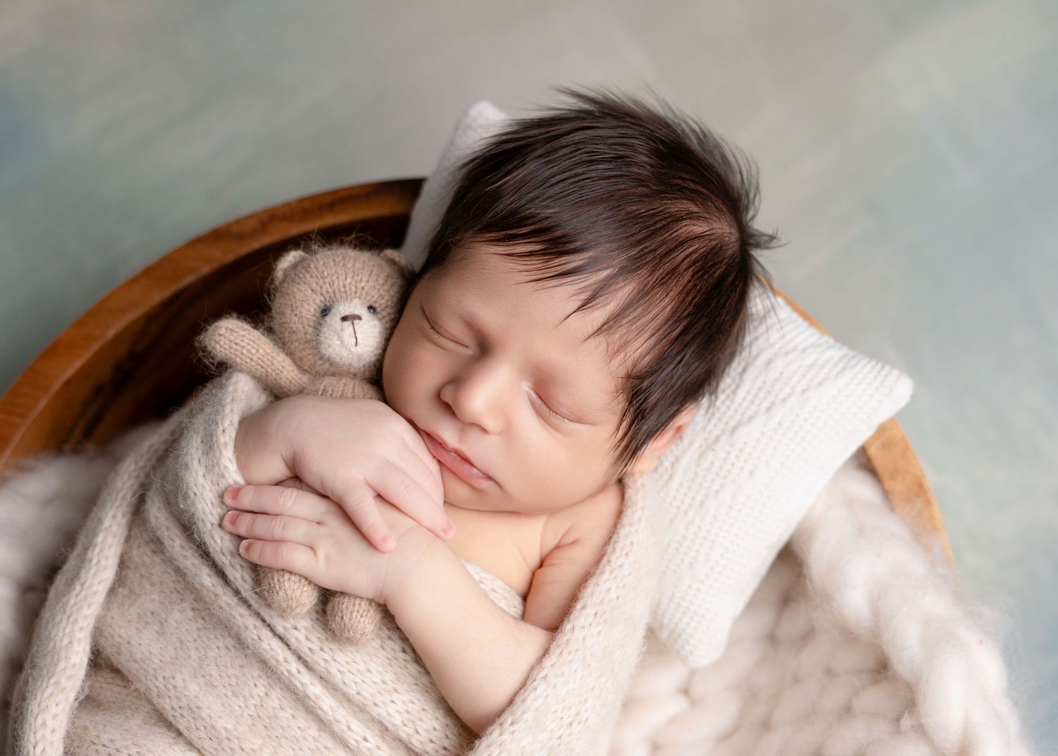 Newborn baby sleeping peacefully in a wooden bowl, wrapped in cream knit blanket, holding a small teddy bear.