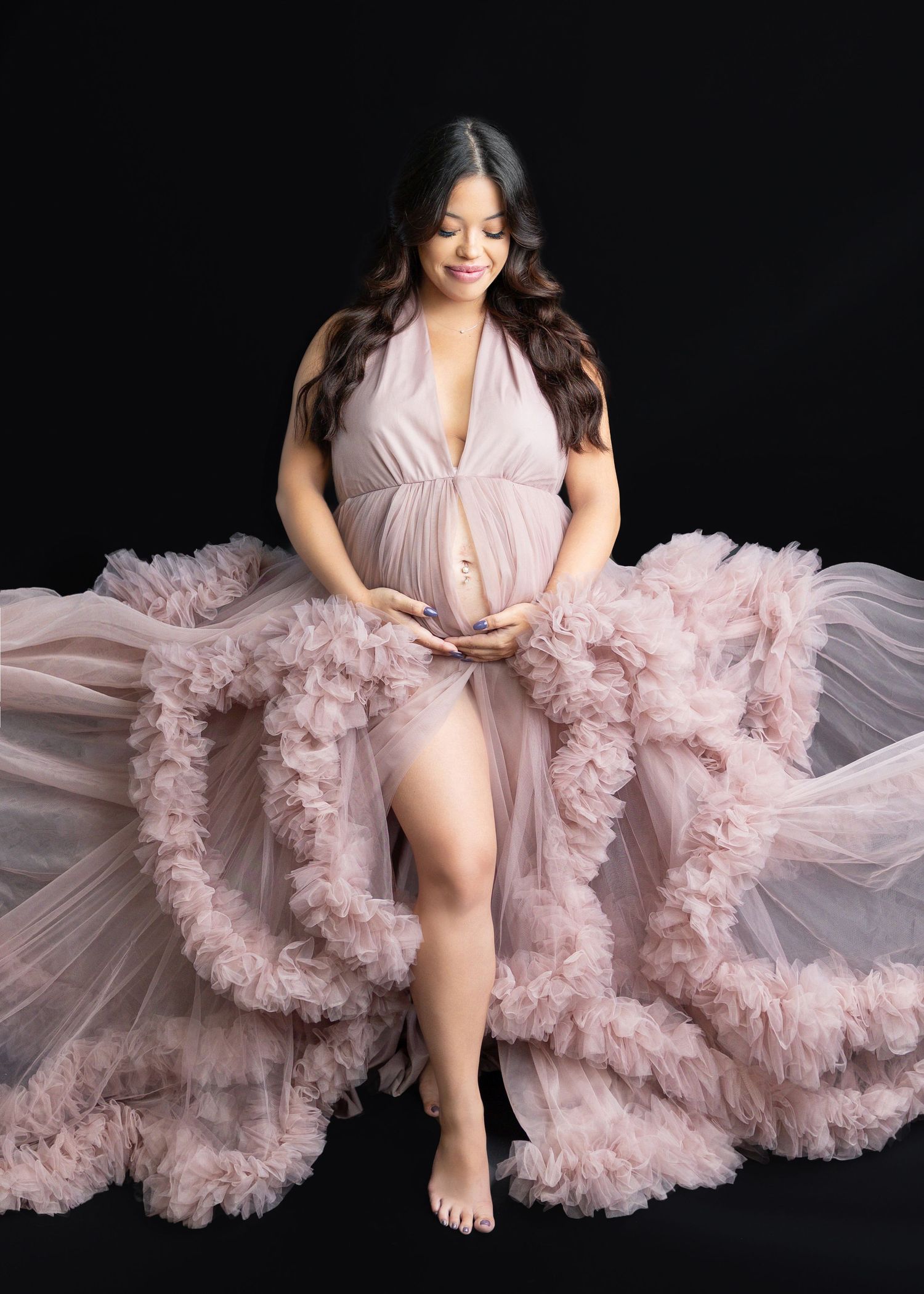 Pregnant woman in flowing pink tulle maternity gown cradling baby bump against black background.