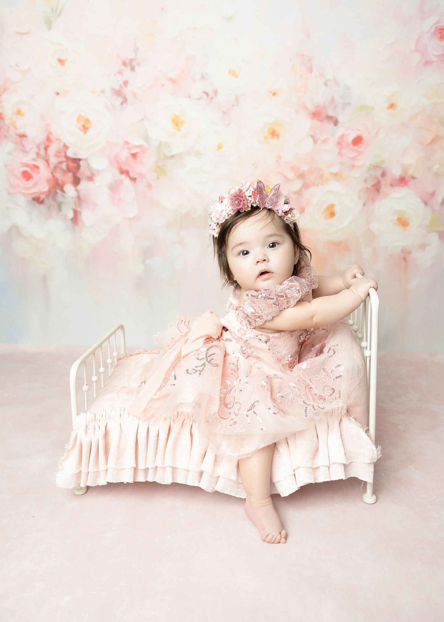Adorable baby in pink embroidered dress and flower crown poses on miniature white bed.