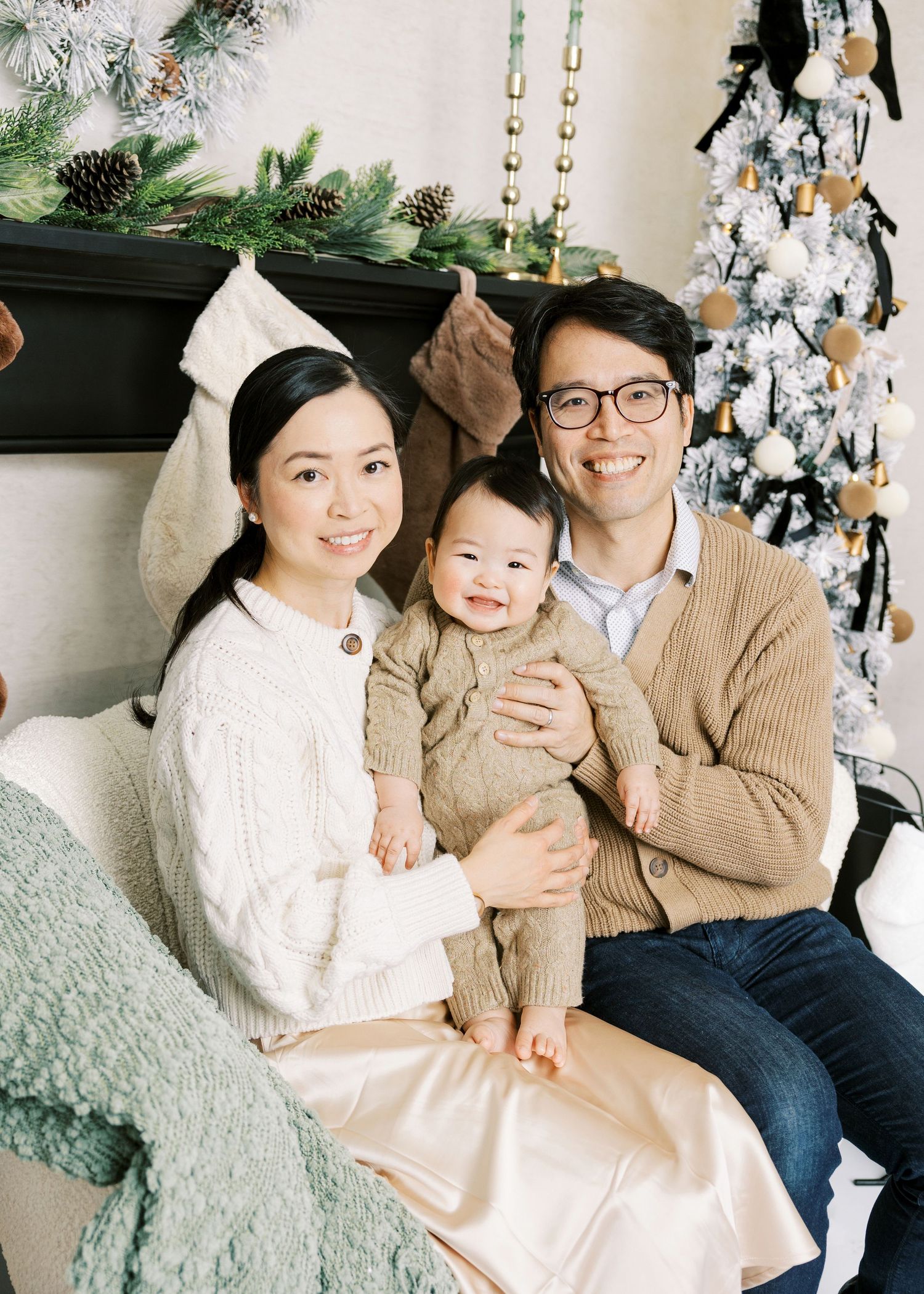Asian family with baby in tan knit outfit posing by Christmas tree and decorated fireplace.