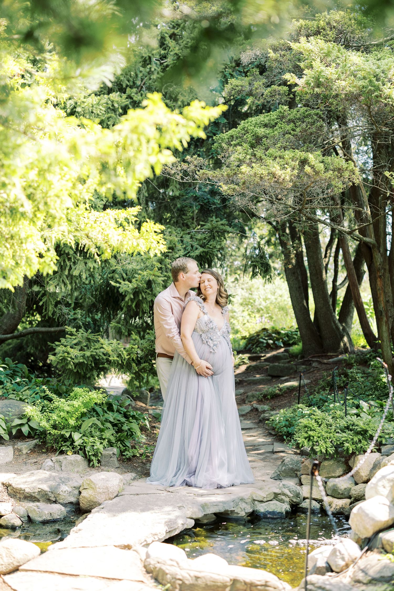 Expecting couple embraces on stone bridge surrounded by lush green trees, woman in lavender maternity gown.