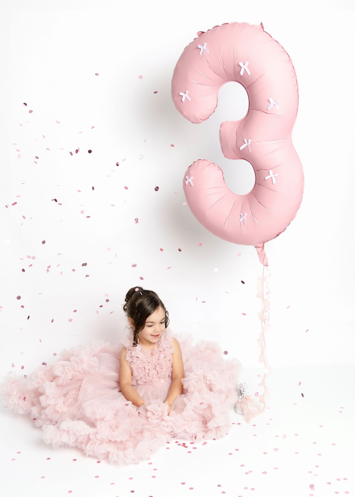 Young girl in pink tulle dress sitting beside a large pink number 3 balloon with confetti falling.