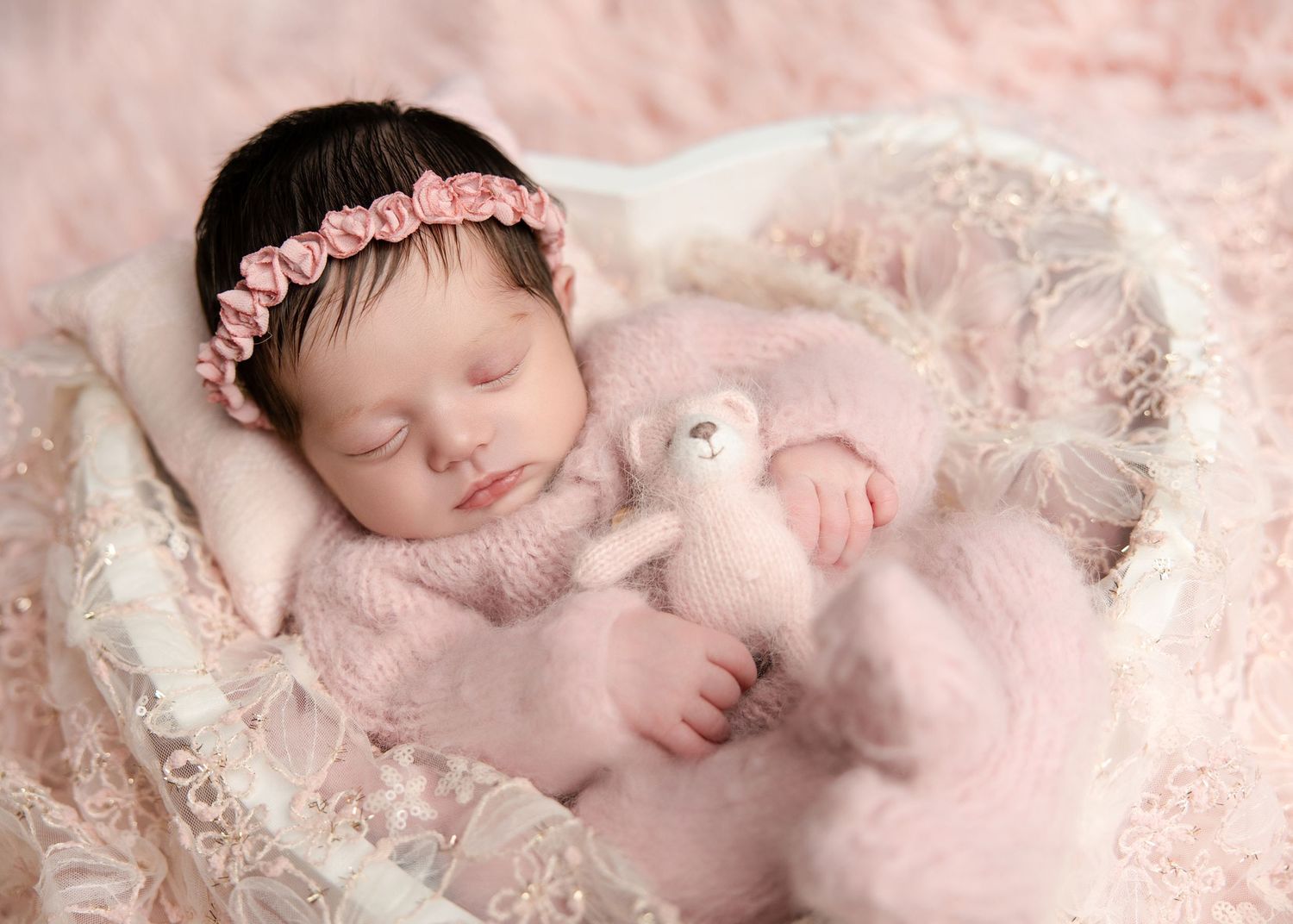 A newborn baby girl in a pink knit outfit sleeps in a basket holding a soft pink teddy bear.