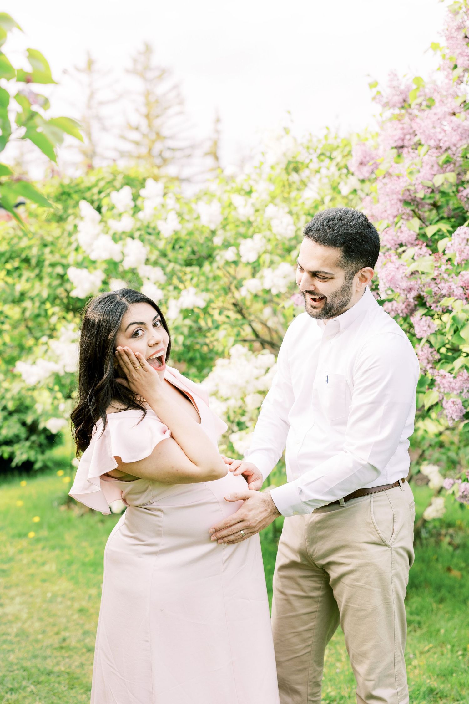 Surprised pregnant woman covers mouth as partner touches her baby bump near blooming white and purple lilacs.