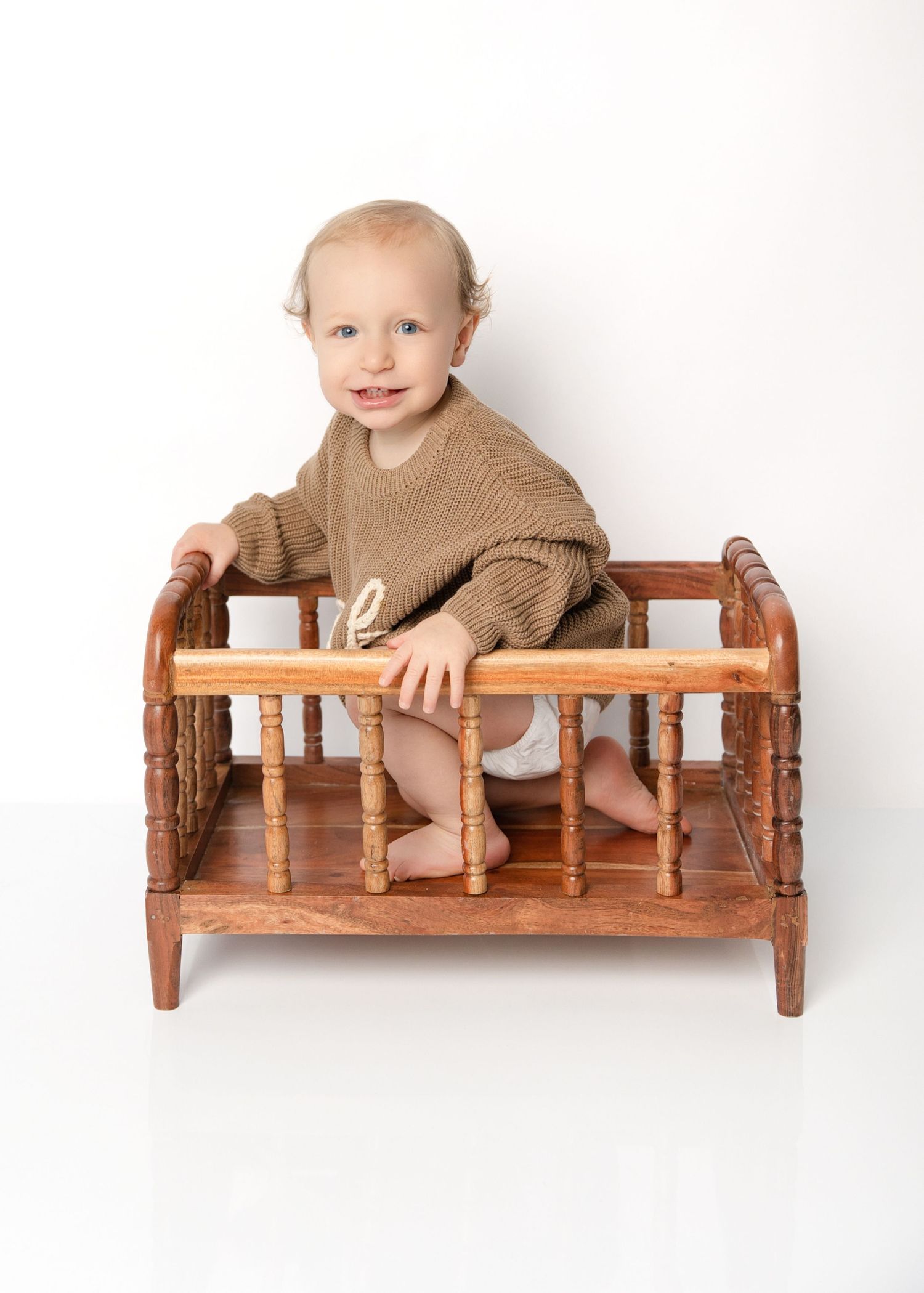 Happy blue-eyed baby in olive sweater sitting inside vintage wooden toy crib on white backdrop.