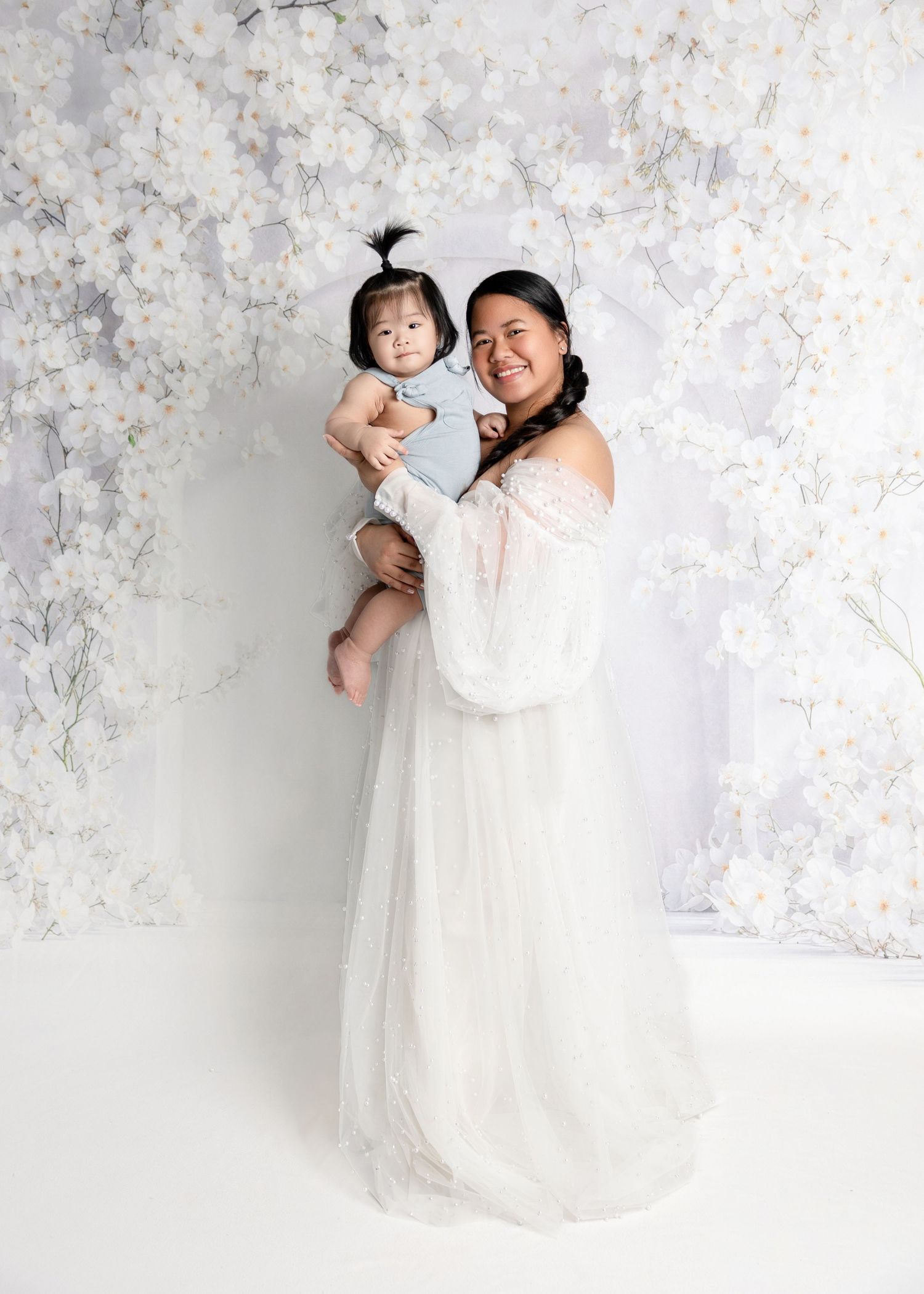 A woman in a white tulle gown holds her baby girl in front of a floral backdrop in a studio portrait.