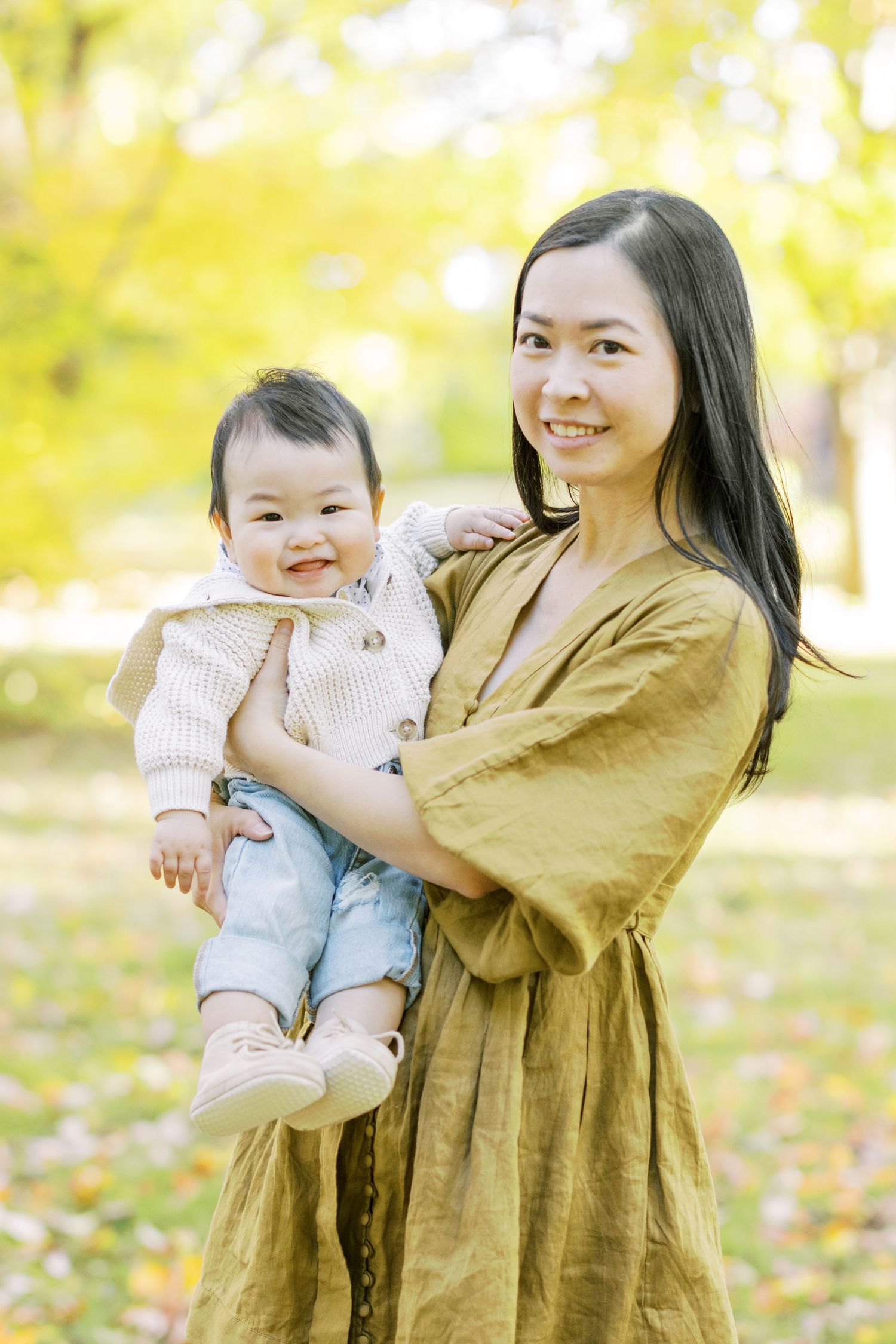 A mother in an olive dress smiles while holding her cheerful baby in a sunny autumn park.
