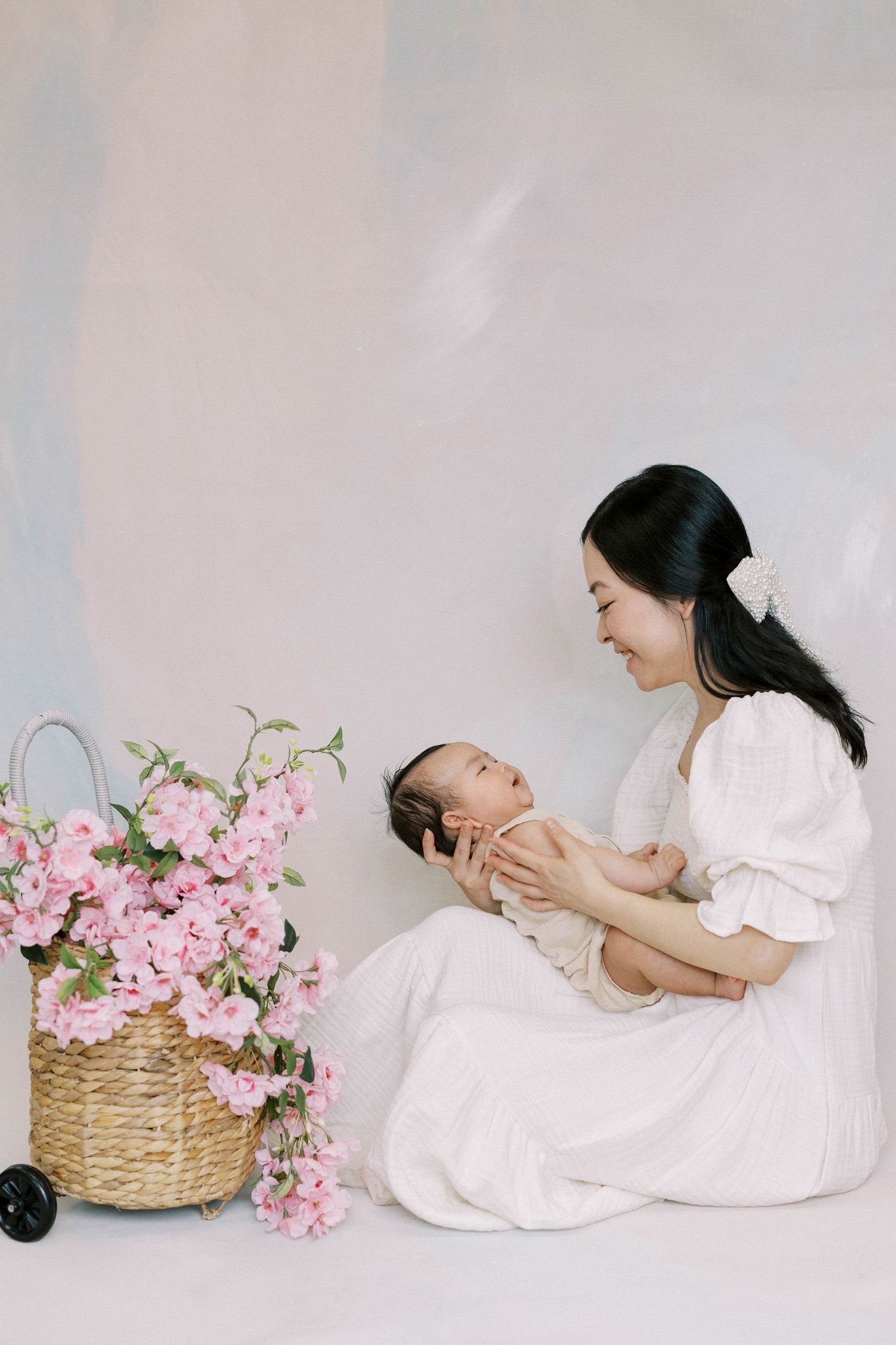 Mother in white dress cradling newborn baby beside basket of pink cherry blossoms in soft studio setting.