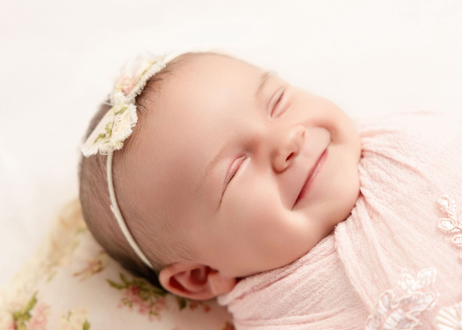 Smiling newborn baby girl swaddled in pink wrap wearing a delicate floral headband.
