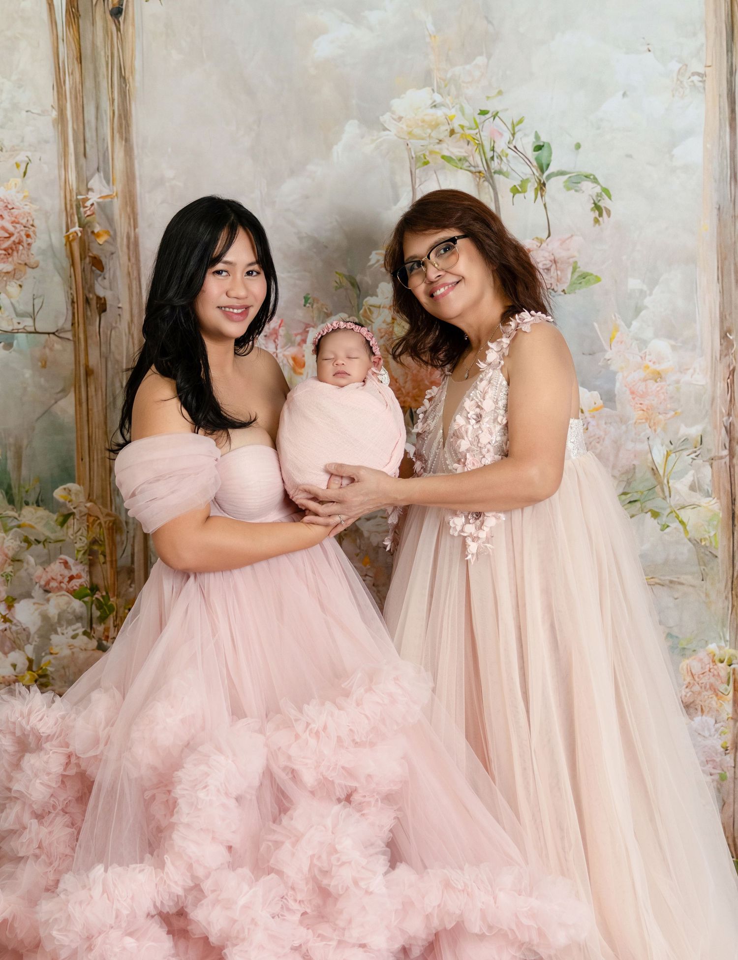 Two women in pink tulle gowns pose with a swaddled newborn baby against a floral backdrop.