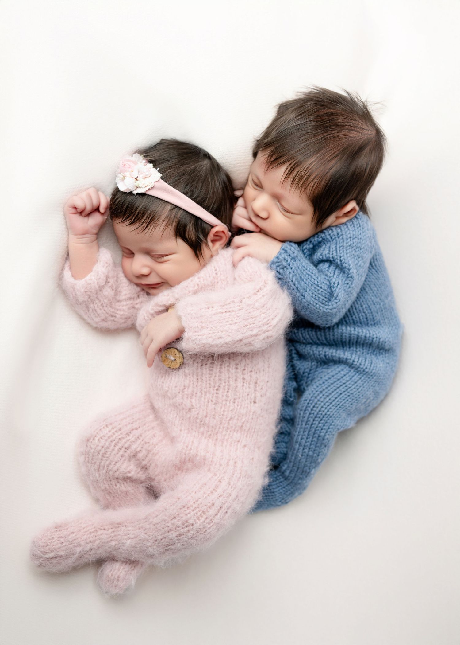 Two sleeping newborn twins, one in pink with a floral headband and one in blue, cuddled together on white background.